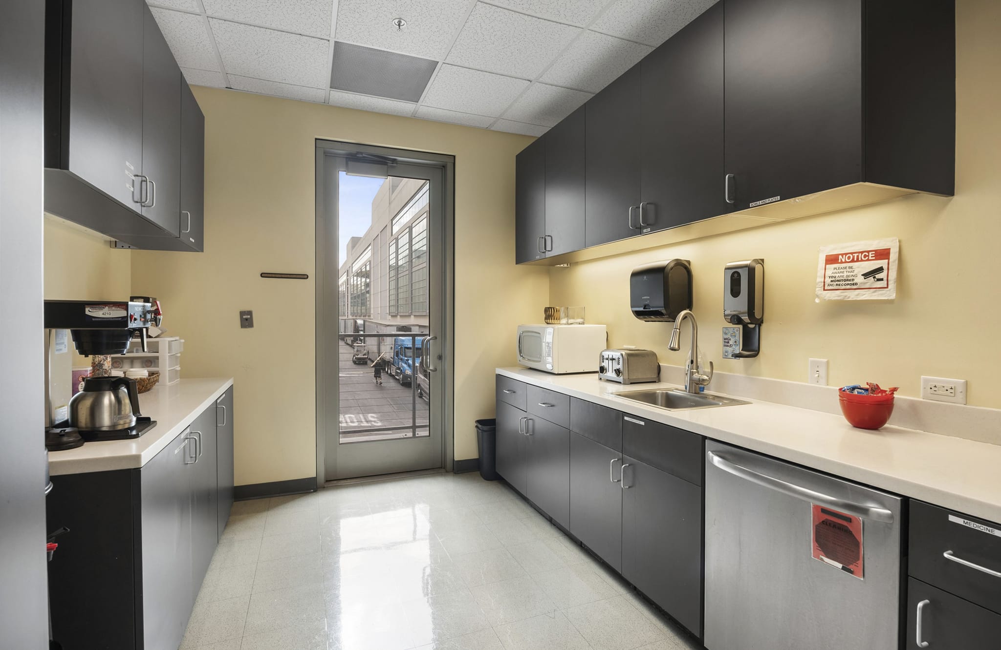 Modern office breakroom with dark gray cabinets, white countertops, appliances, and a glass door.