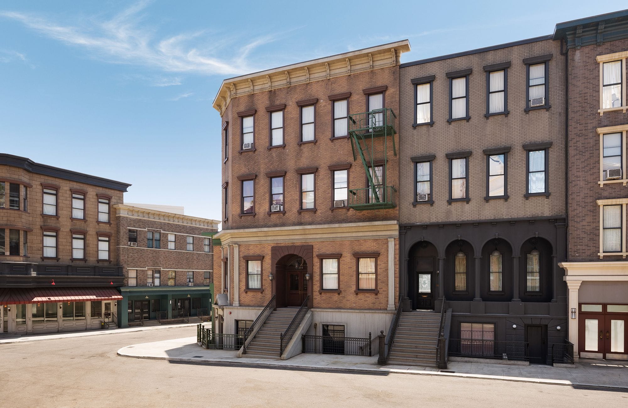 A street corner with multi-story urban buildings, one featuring a green fire escape, under a blue sky.