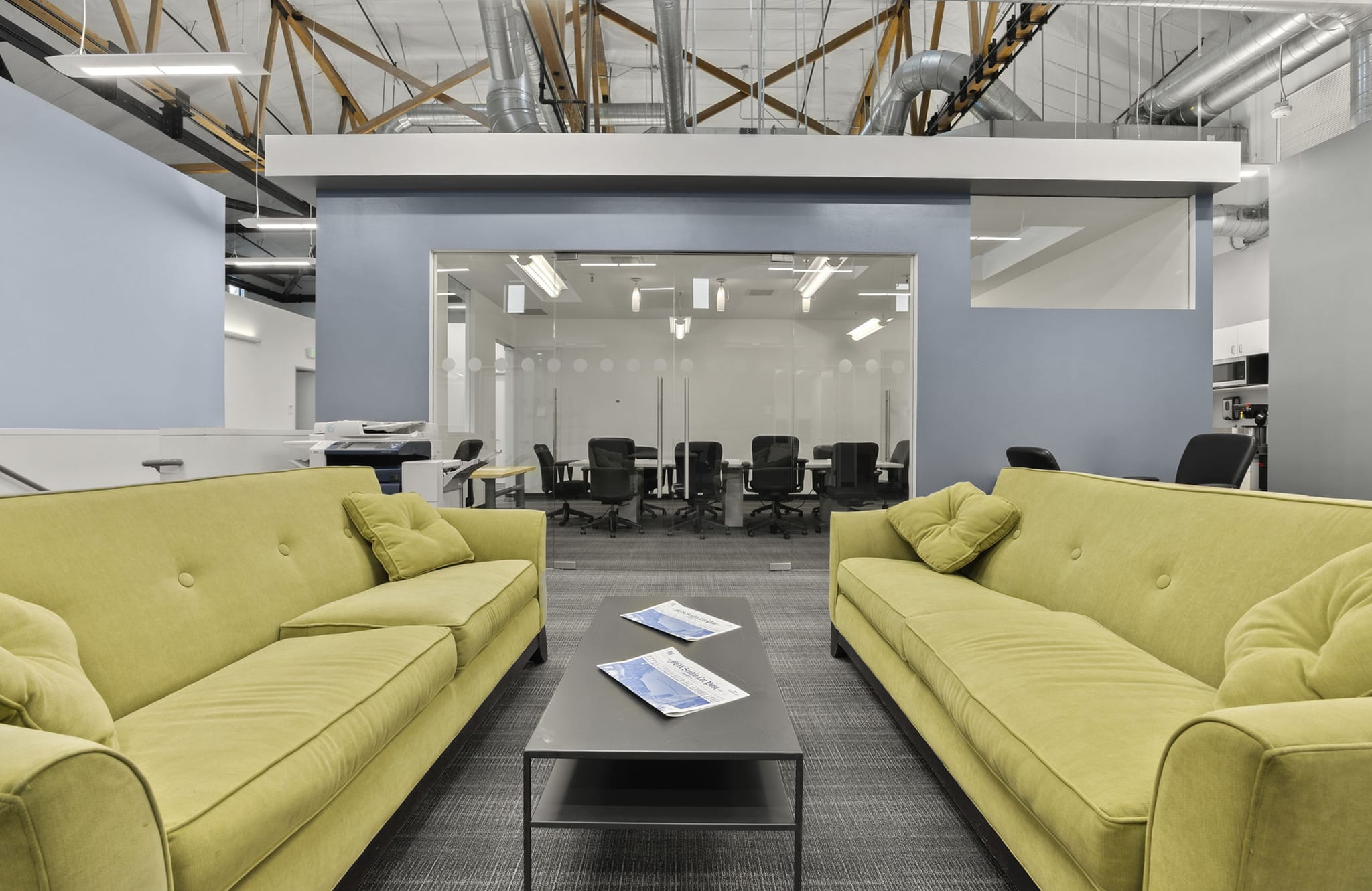 Modern office lounge with two chartreuse couches, a coffee table, and a conference room, under an exposed ceiling.