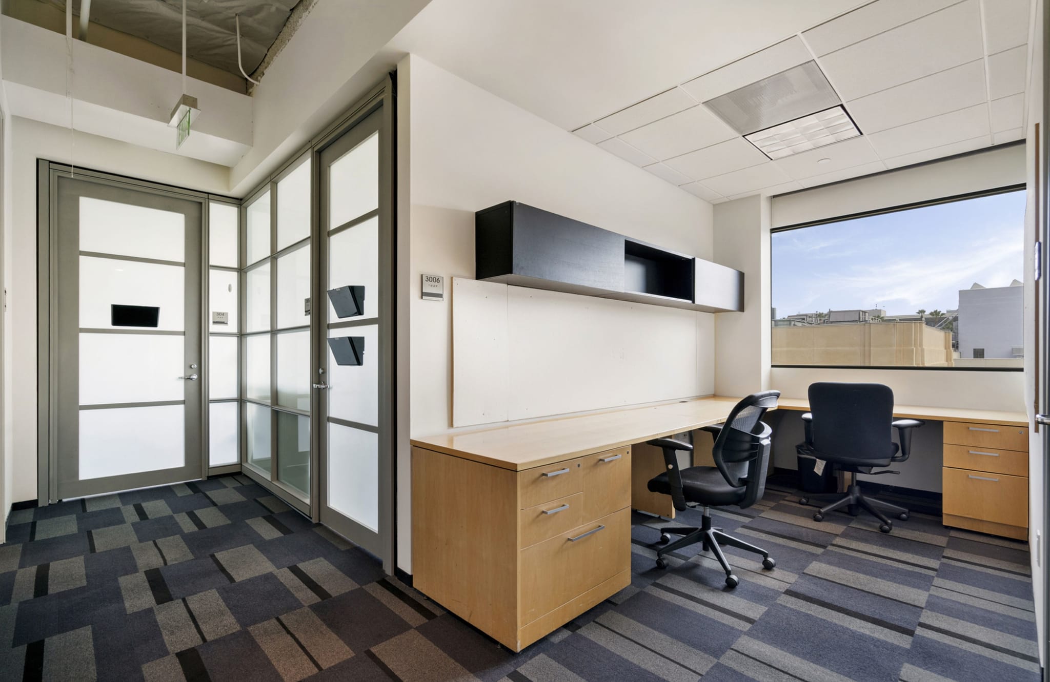An empty office with two desks, two black office chairs, black overhead shelving, a large window, and a frosted glass partition.
