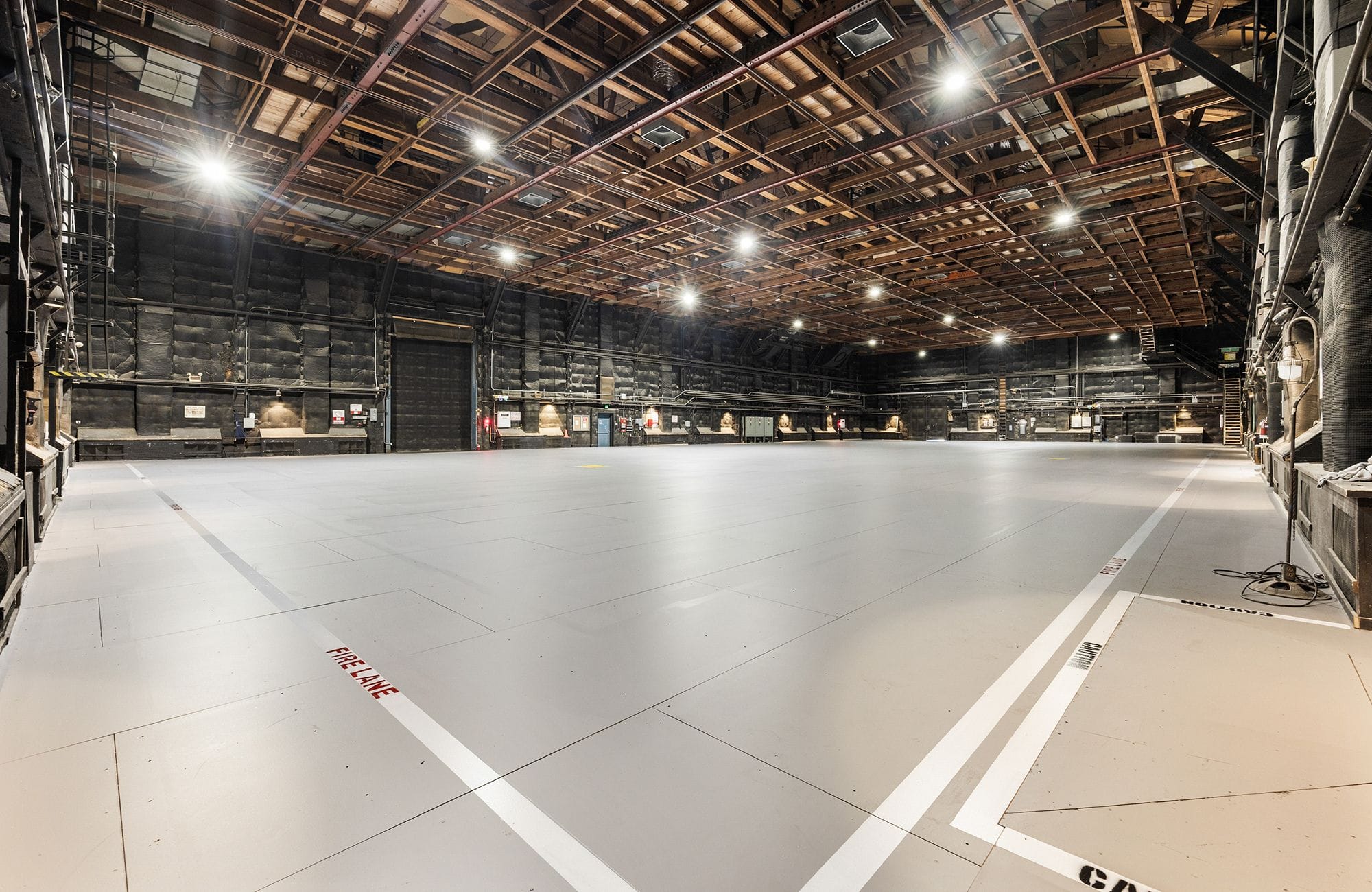 An empty soundstage with a light grey floor, dark walls, exposed wooden ceiling, and bright overhead lights.