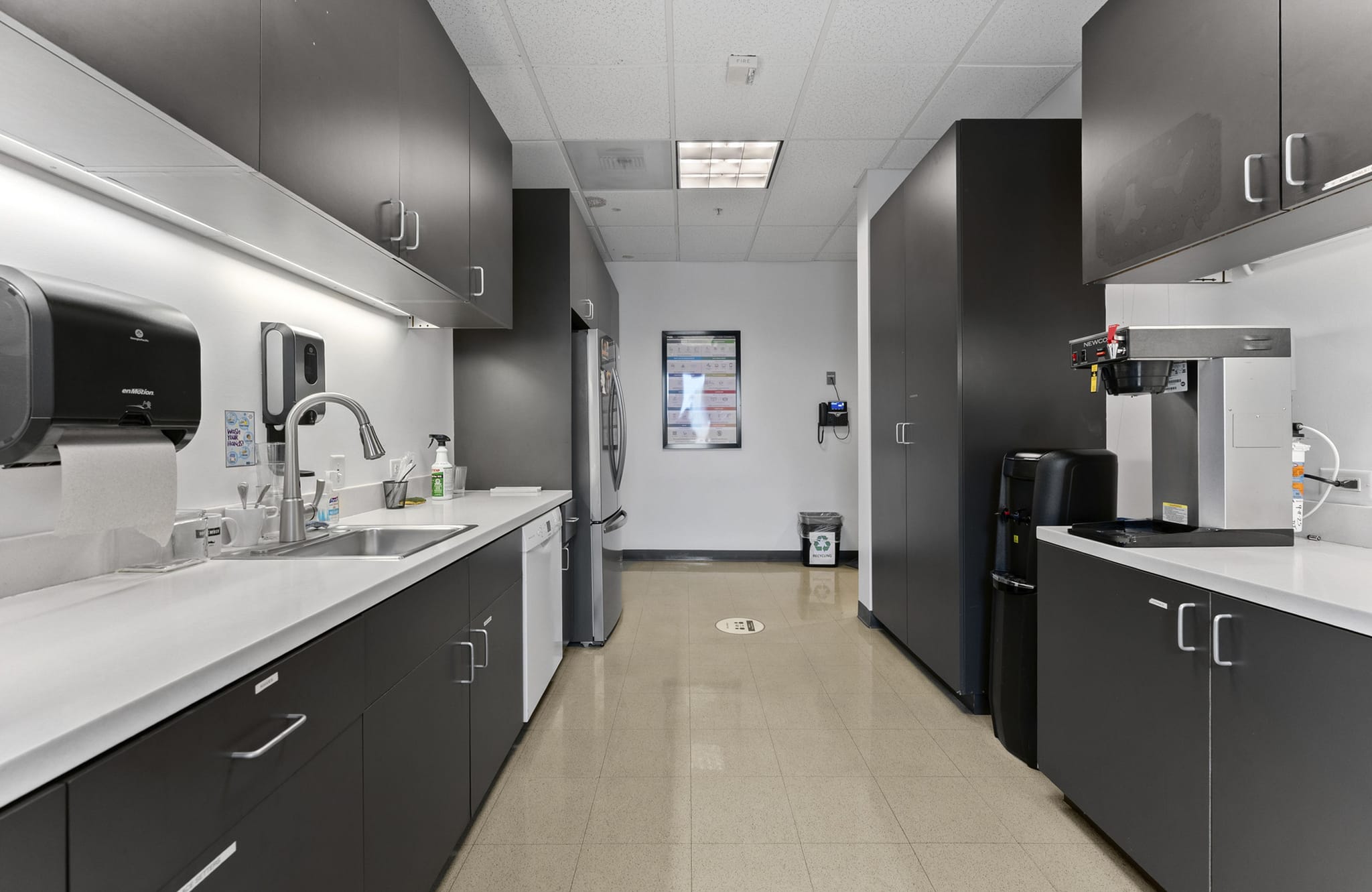 Modern office breakroom with dark gray cabinets, white countertops, a sink, and a coffee machine.