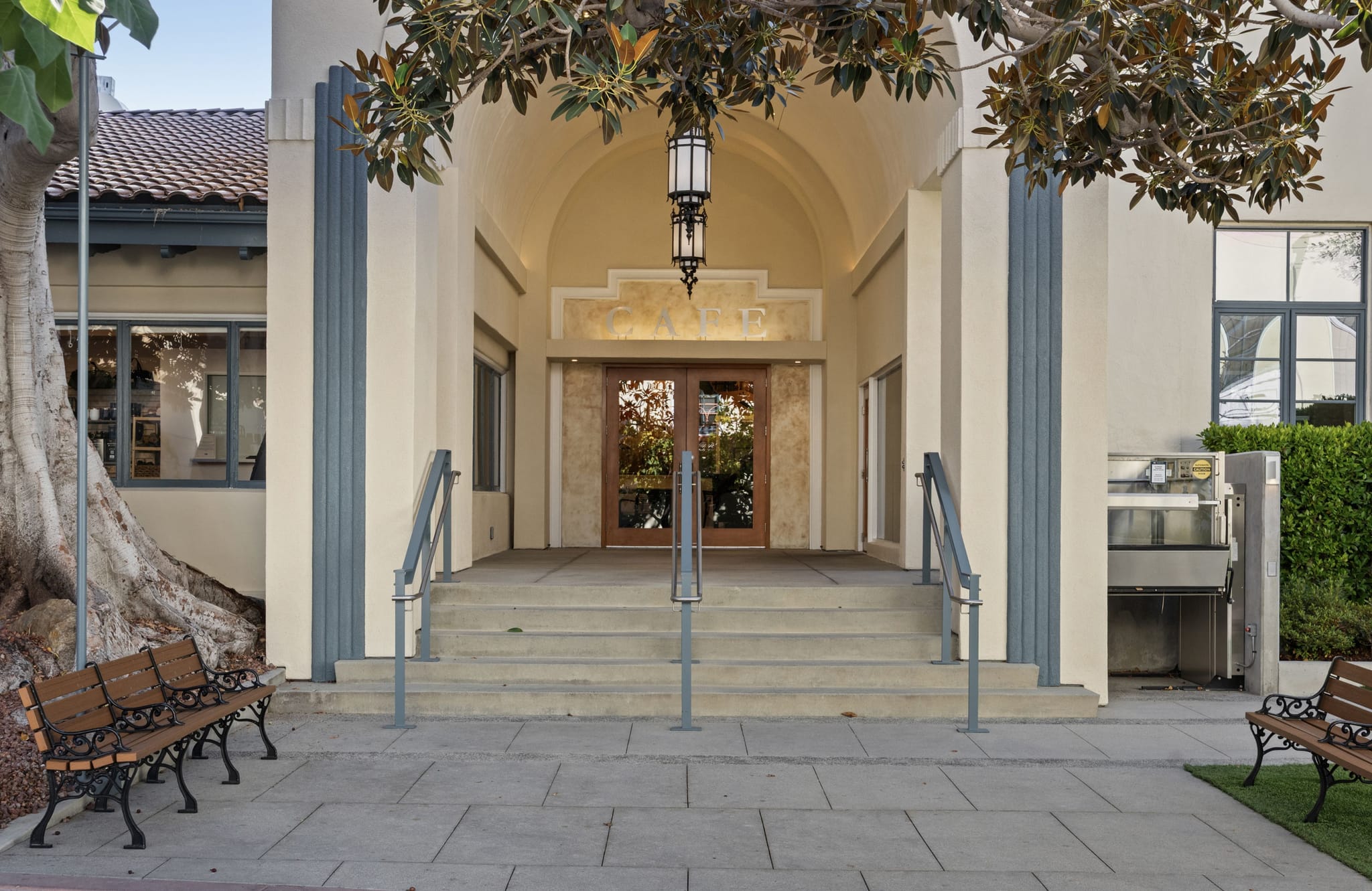 Building entrance with steps, an arched doorway, a "CAMP" sign, and an accessibility lift to the right, flanked by benches and trees.