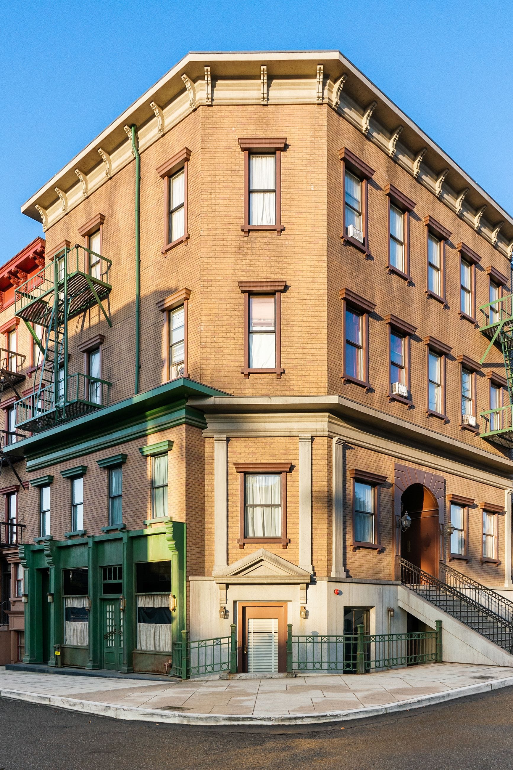 A brown brick corner building with green storefronts and fire escapes under a blue sky.