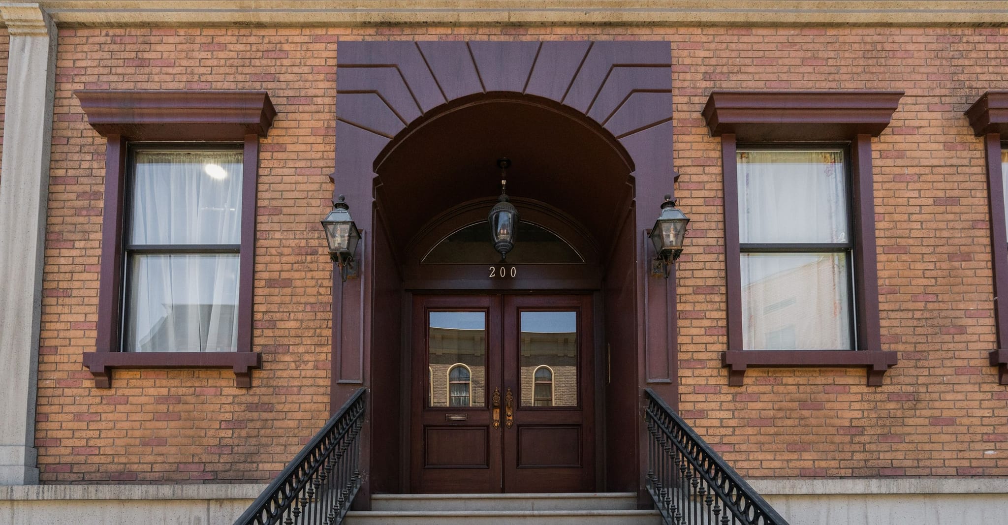Front entrance of a brick building with dark brown double doors numbered "200" under an arch, flanked by windows and railings.