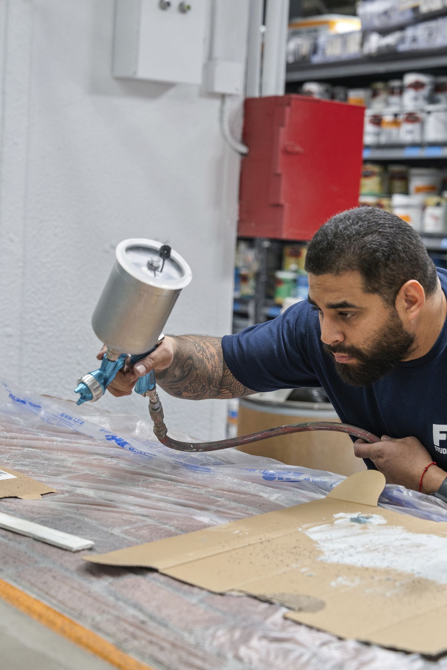 A man with a beard and tattoos on his arm operates a paint spray gun over a covered work surface.