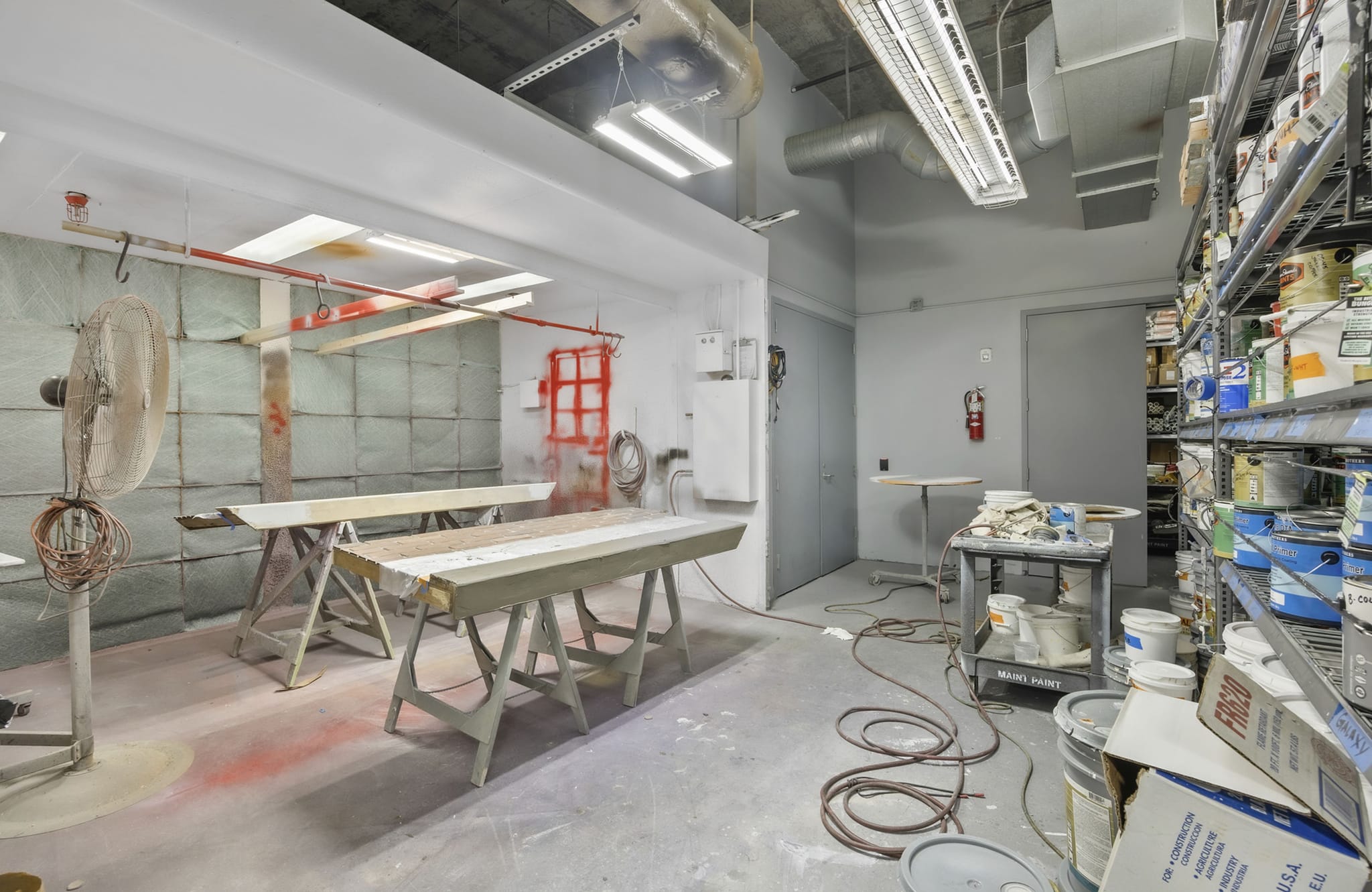 An industrial workshop with a spray painting booth on the left and shelves of paint supplies on the right.