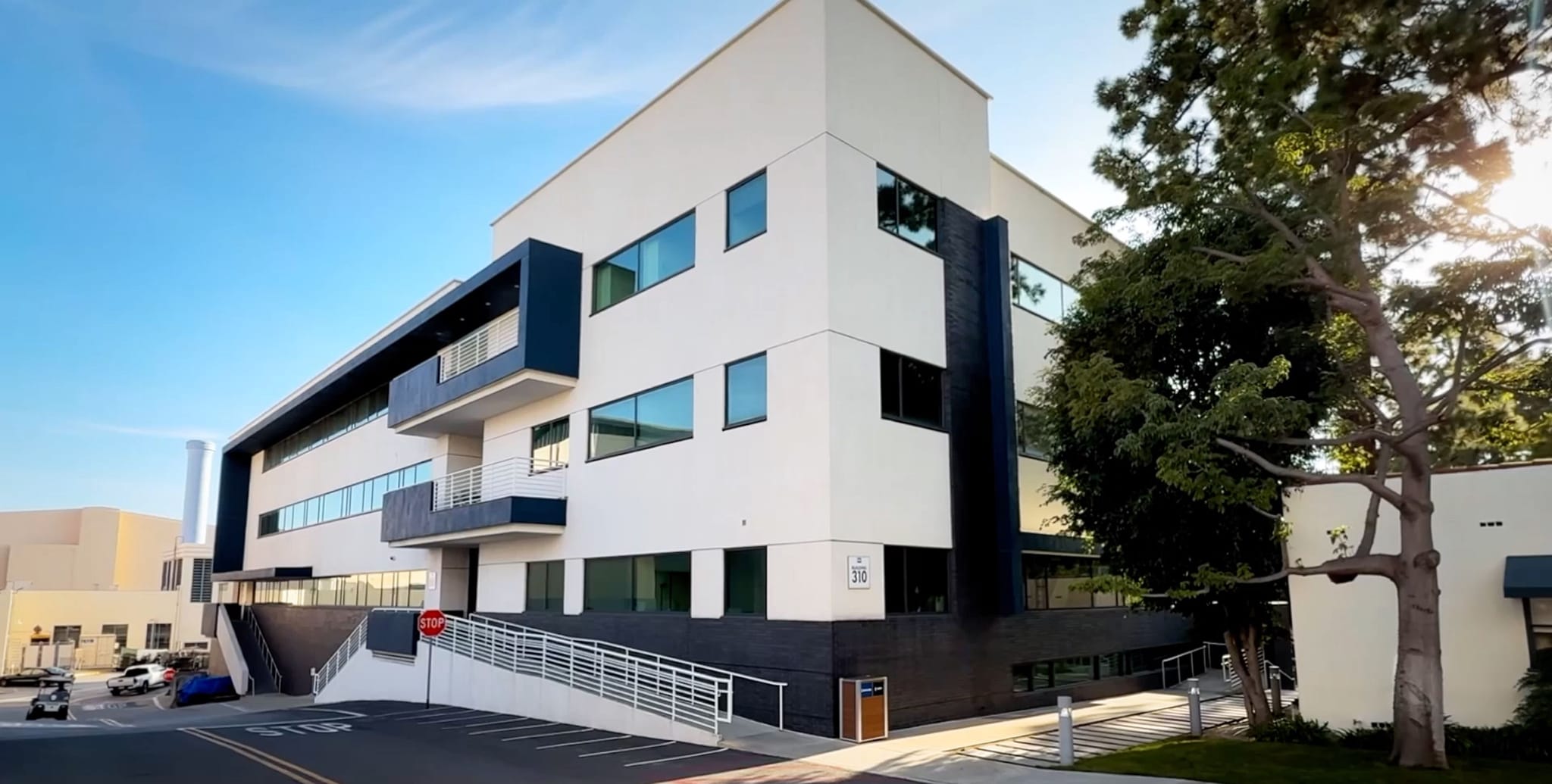 A modern multi-story building with white and dark blue sections and many windows, under a clear blue sky, with a large tree to the right.