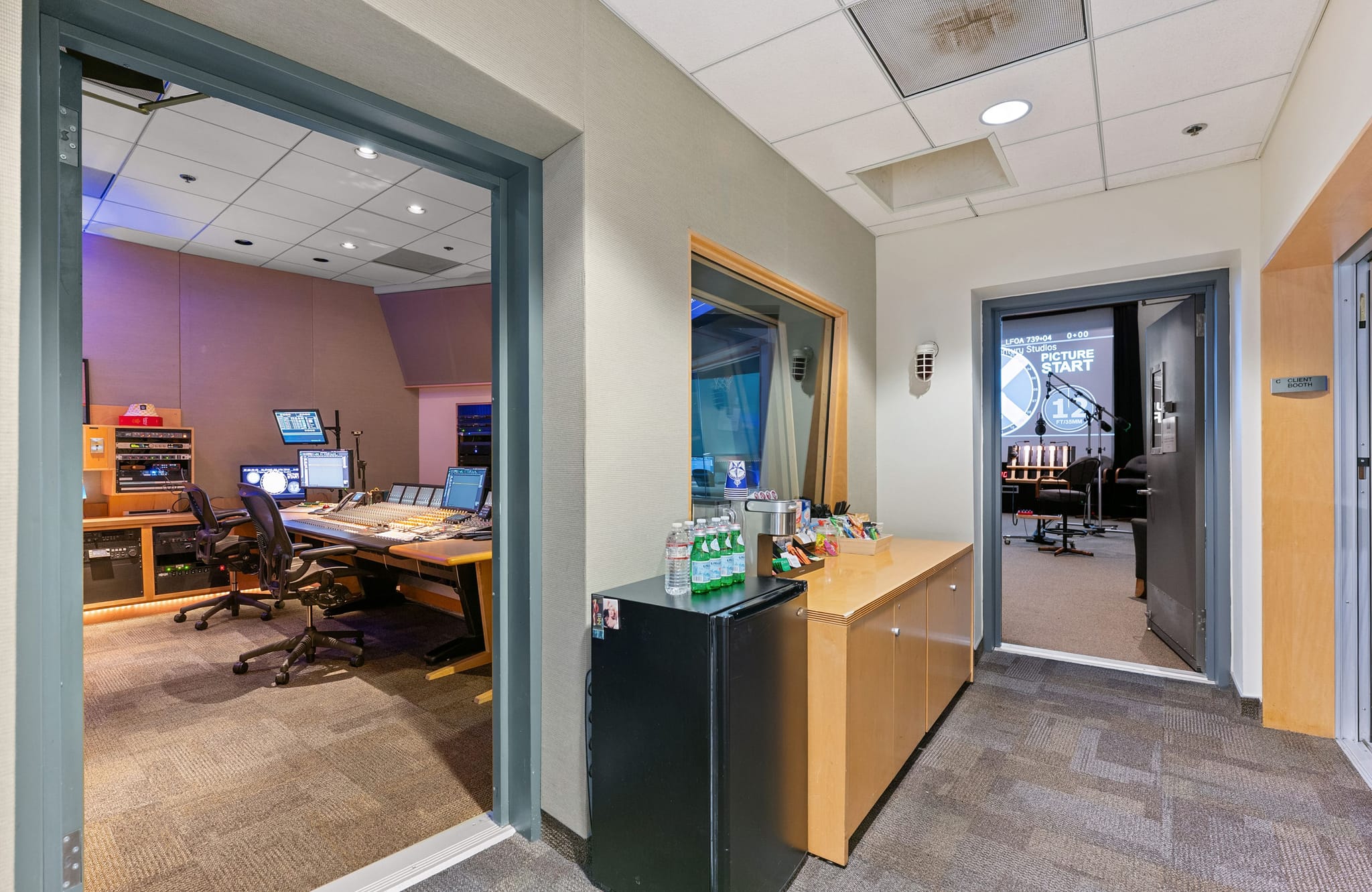 A hallway connecting a multi-monitor control room to the left and a recording studio to the right, with a snack and drink counter in between.