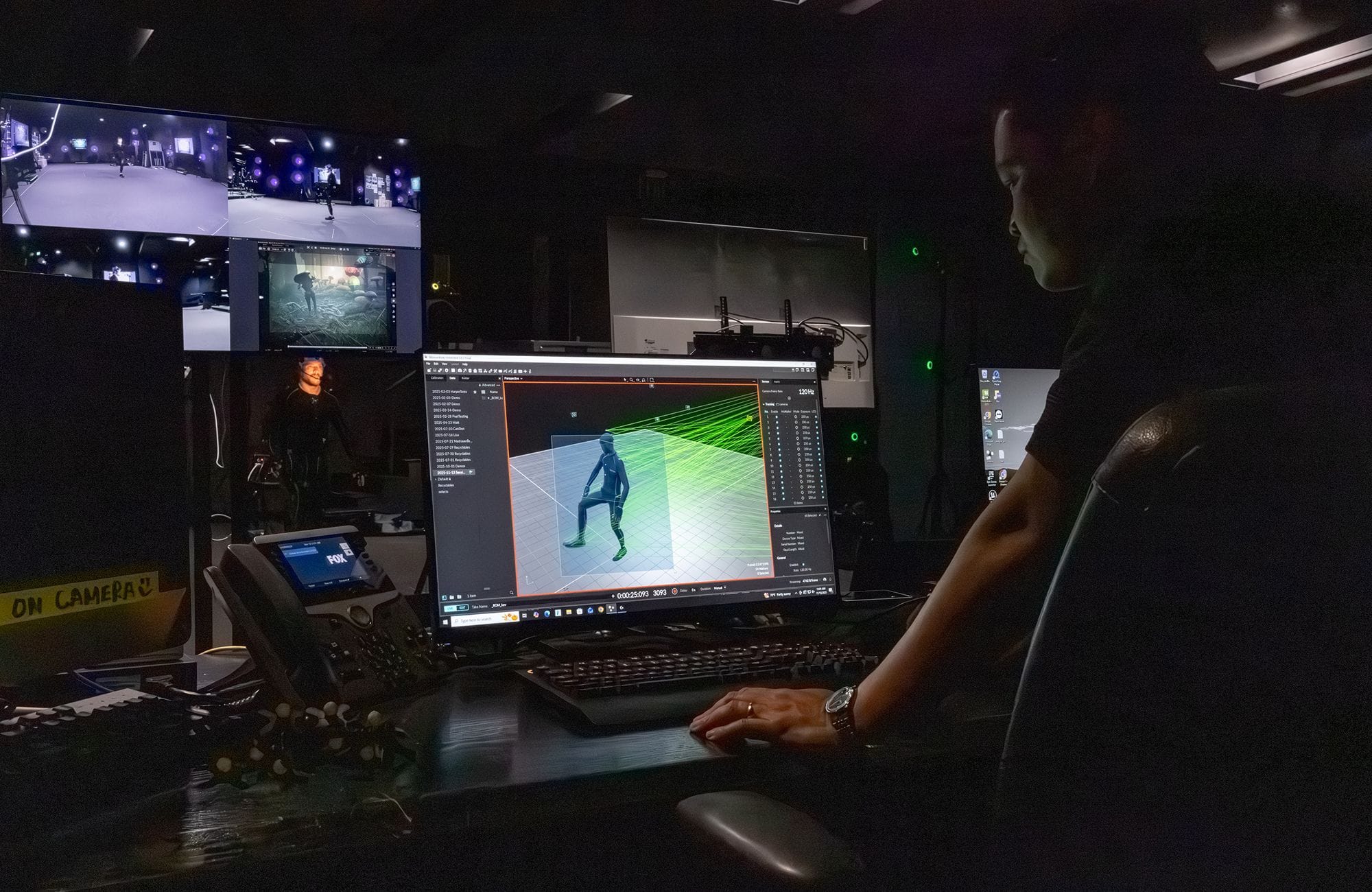 A person works on a computer in a dark control room, monitoring a 3D motion-capture figure on screen, with live studio feeds in the background.