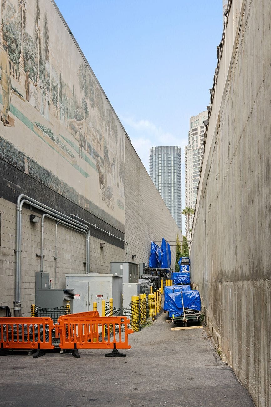 A narrow urban alley with a faded mural on the left wall and a plain concrete wall on the right, leading towards distant skyscrapers. Orange barriers and blue-tarped items are visible on the ground.
