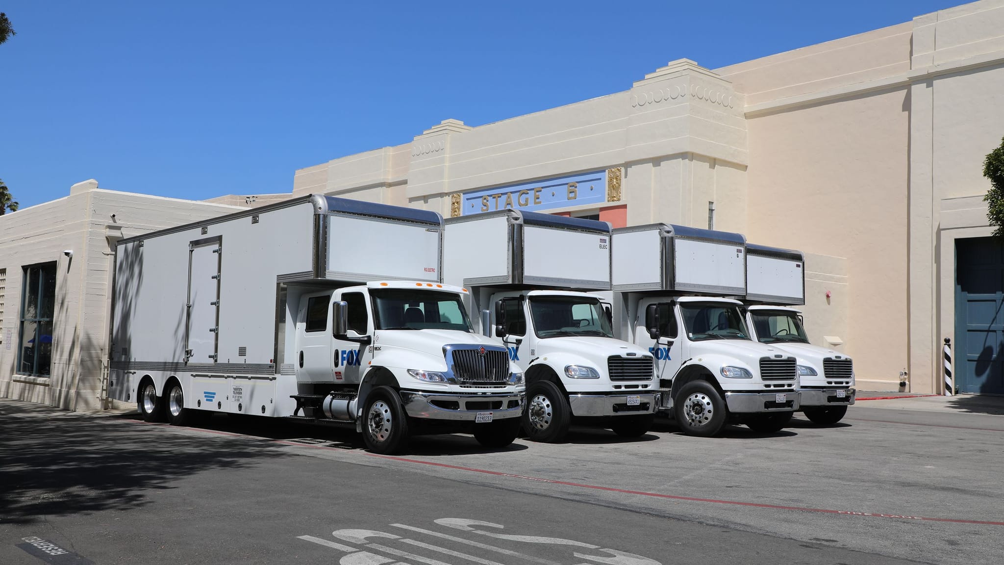 Four white film production trucks parked in front of a studio building labeled "STAGE B".