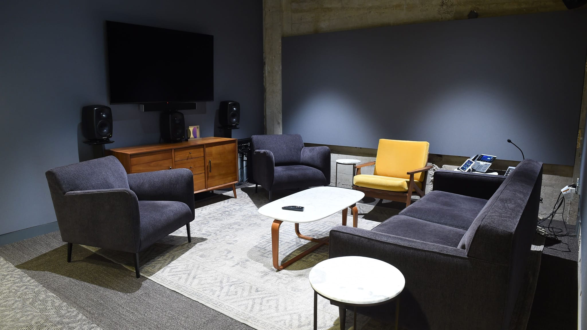 A contemporary living room with a large wall-mounted TV, a wooden credenza, a dark gray sofa, two dark gray armchairs, a yellow armchair, and a coffee table on a patterned rug.