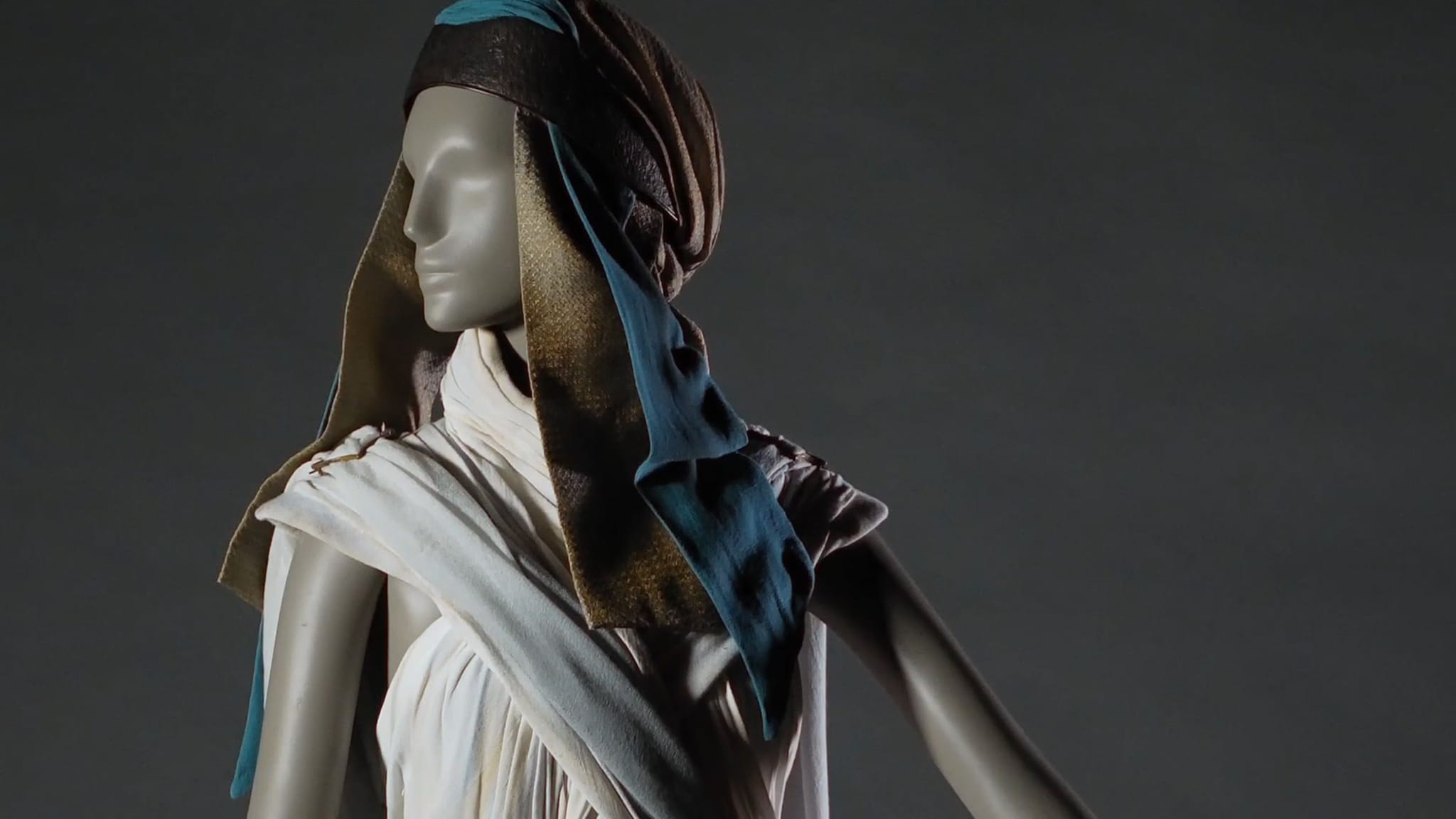 Mannequin in a white pleated garment with a brown and blue patterned headpiece.
