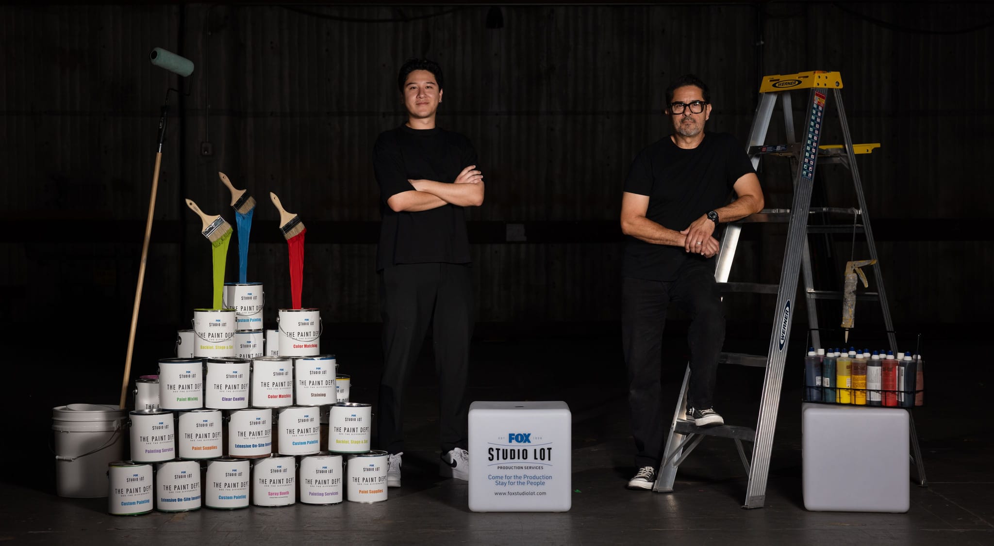 Two men in a dark studio pose with painting supplies, including a pyramid of paint cans, colorful brushes, a ladder, and a FOX STUDIO LOT branded box.