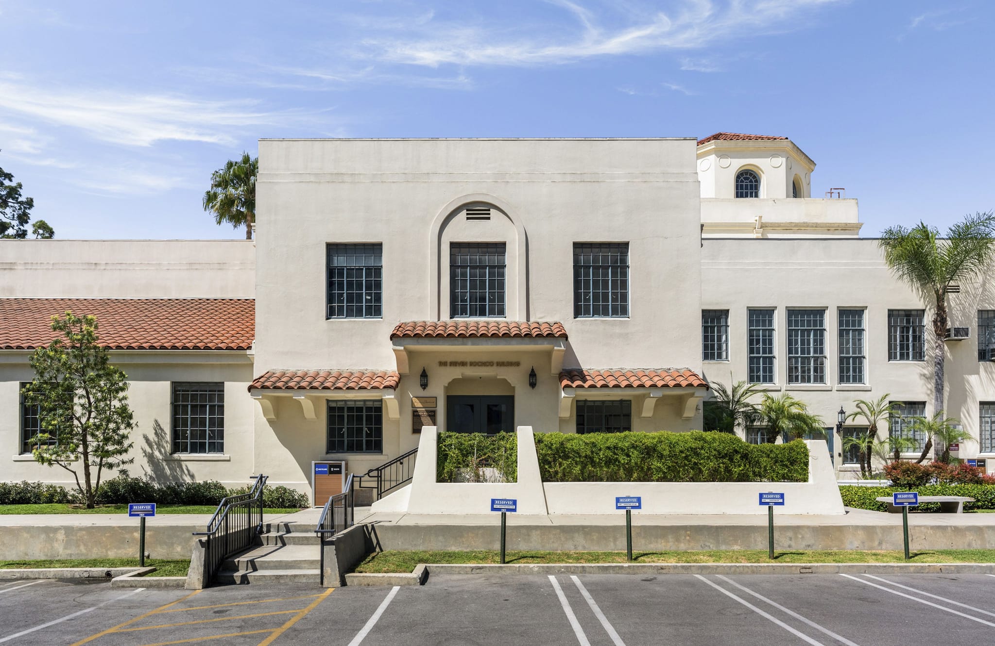 A light-colored, Mediterranean-style building with a red tile roof and an arched entrance, surrounded by landscaping, with a parking lot in the foreground.