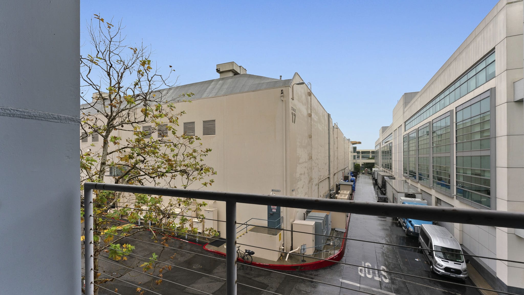 View from a balcony railing of a wet street between large commercial buildings and a tree, with parked vans.