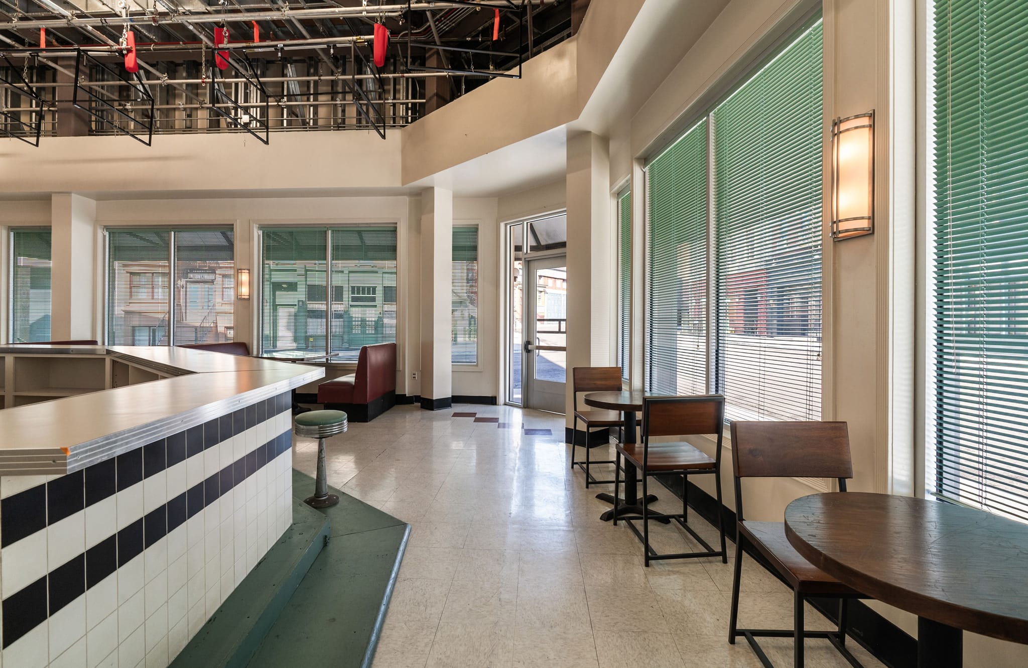 An empty retro-style diner interior with a black and white counter, green bar stool, red booths, and cafe tables near windows with green blinds.