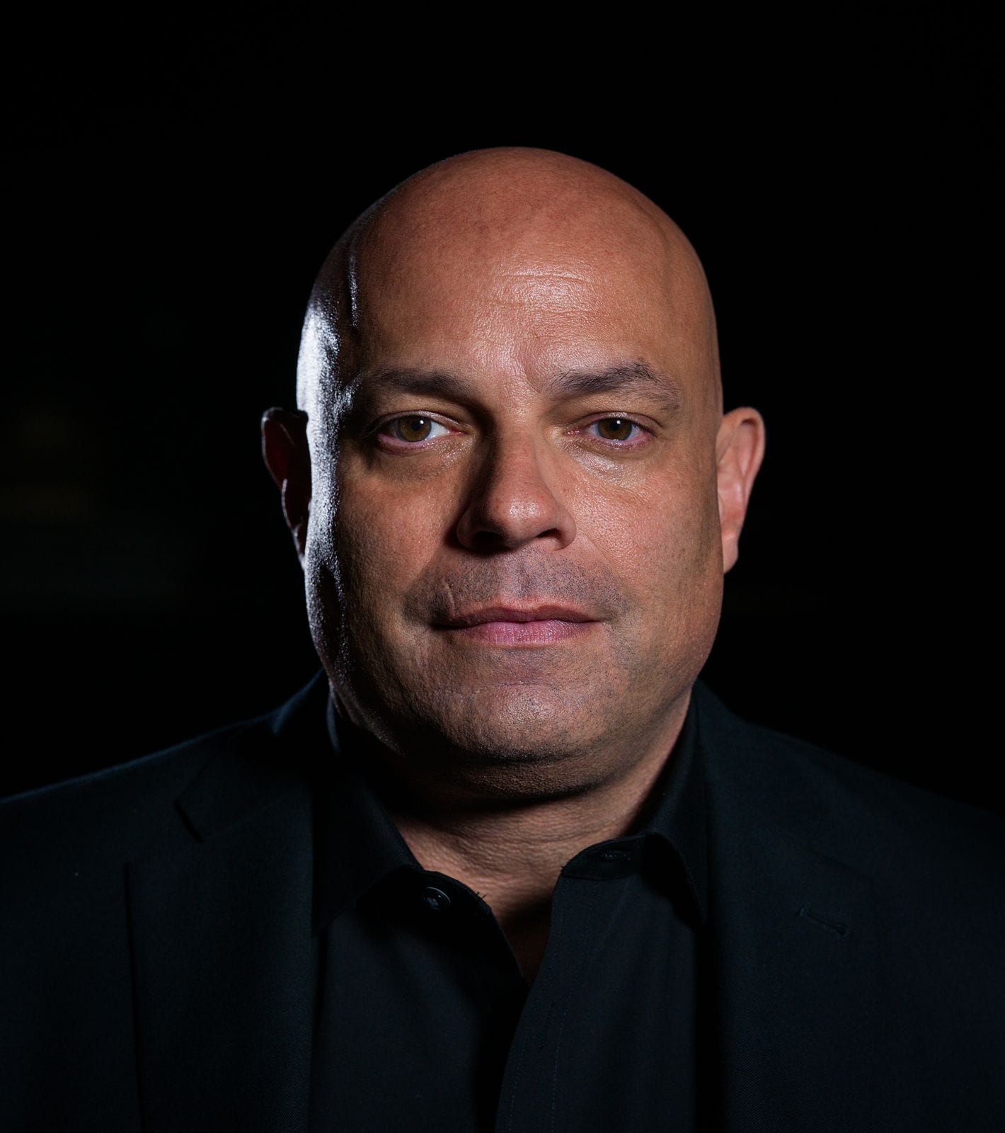 A bald man in a black shirt and jacket looks directly at the camera against a dark background.