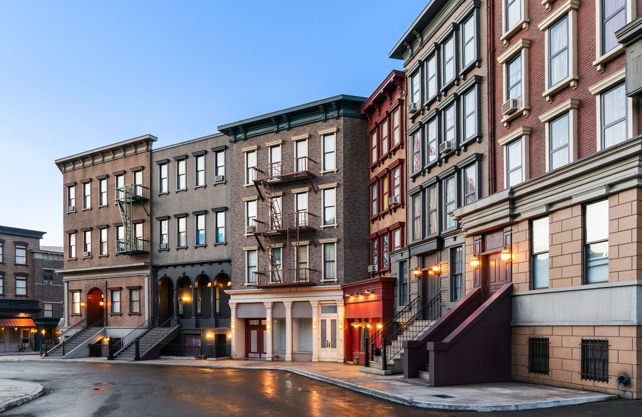A row of old city buildings with fire escapes on a wet street at dusk, with streetlights lit.