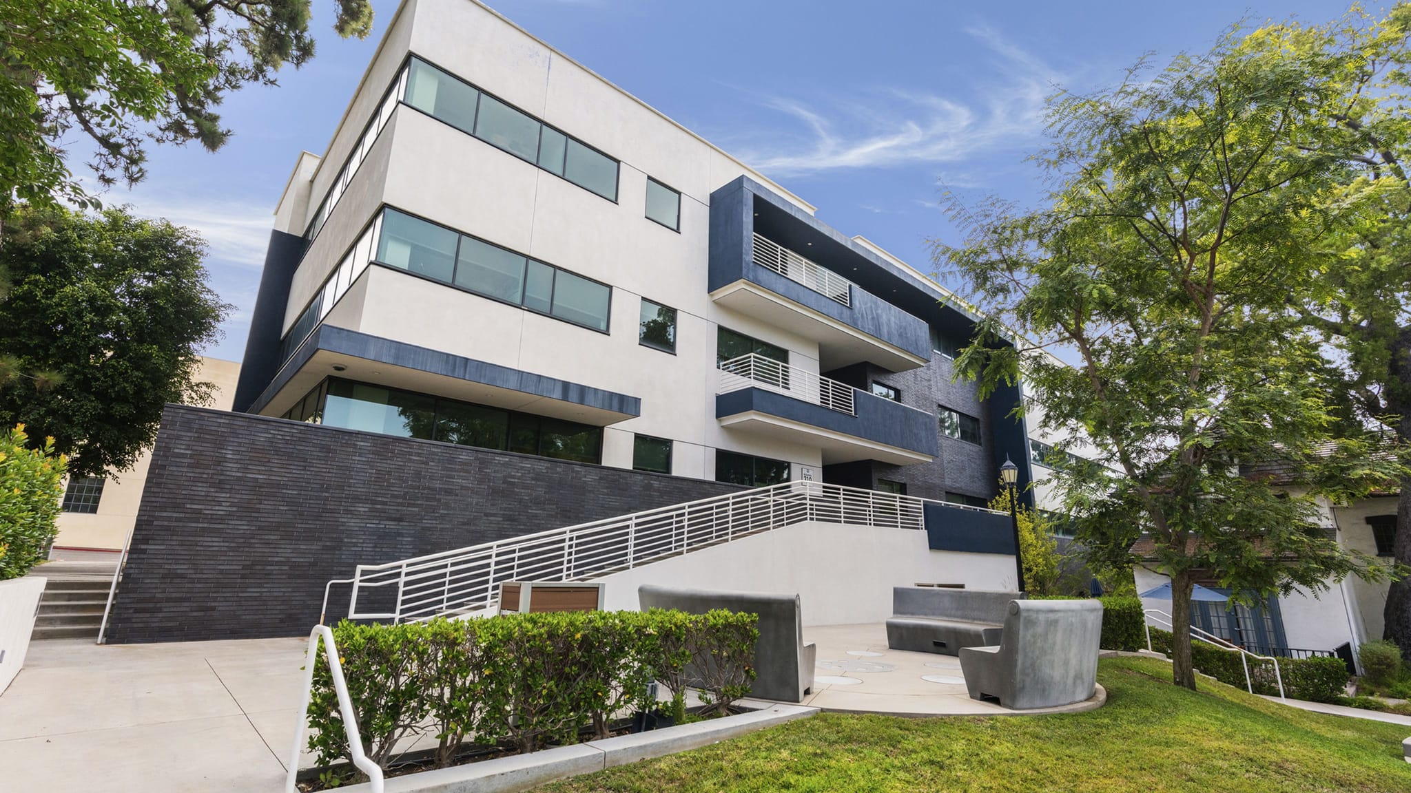 A modern white and dark blue building with large windows, balconies, and an outdoor staircase, surrounded by green landscaping.