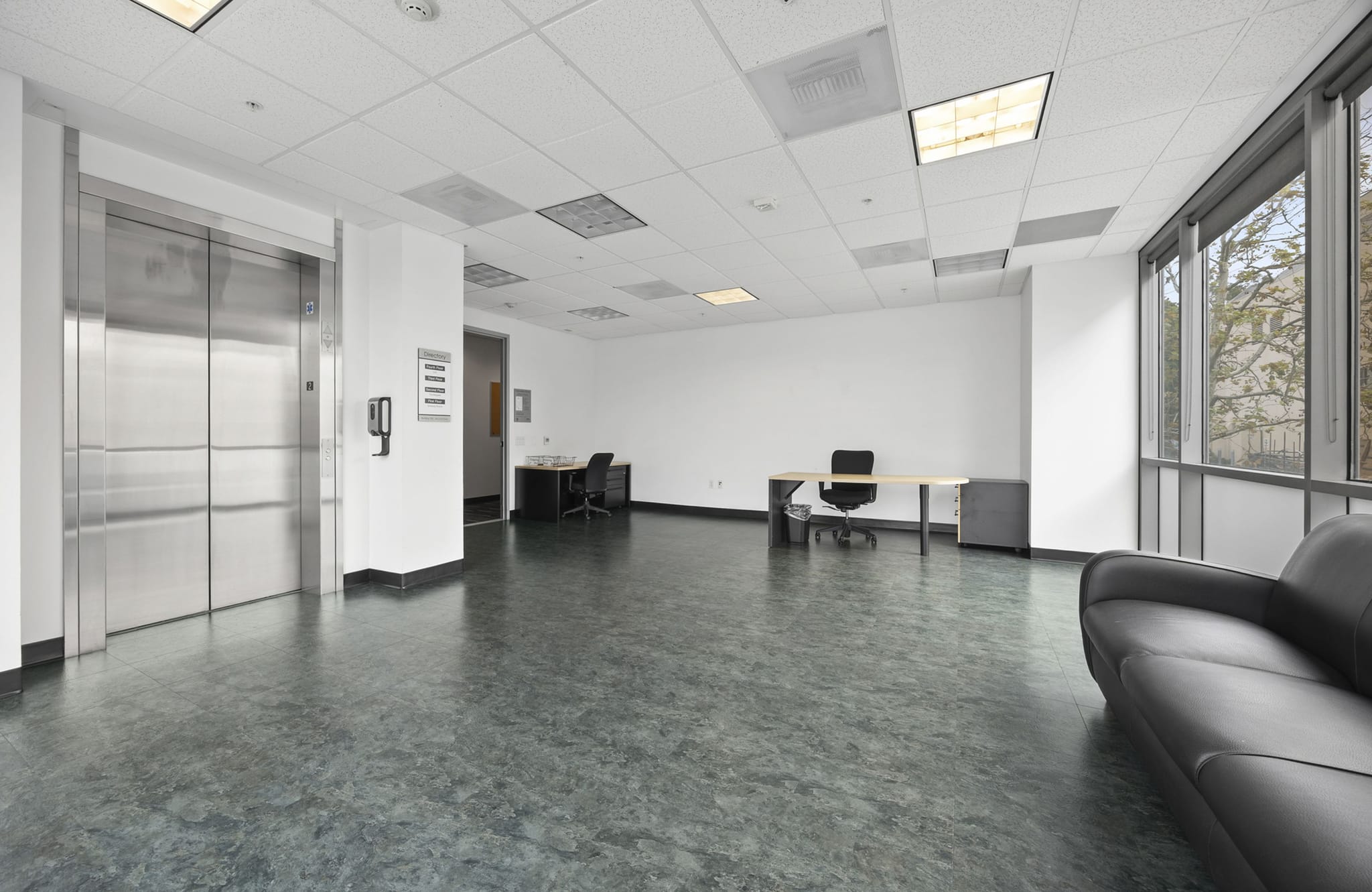 Commercial space with a stainless steel elevator, large windows, two desks, and a dark sofa on a grey floor.