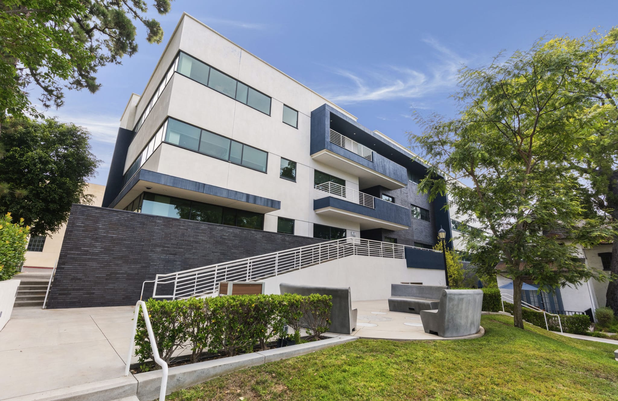 A modern white and dark blue building with balconies, a black stone lower section, and a white staircase, surrounded by green landscaping under a blue sky.