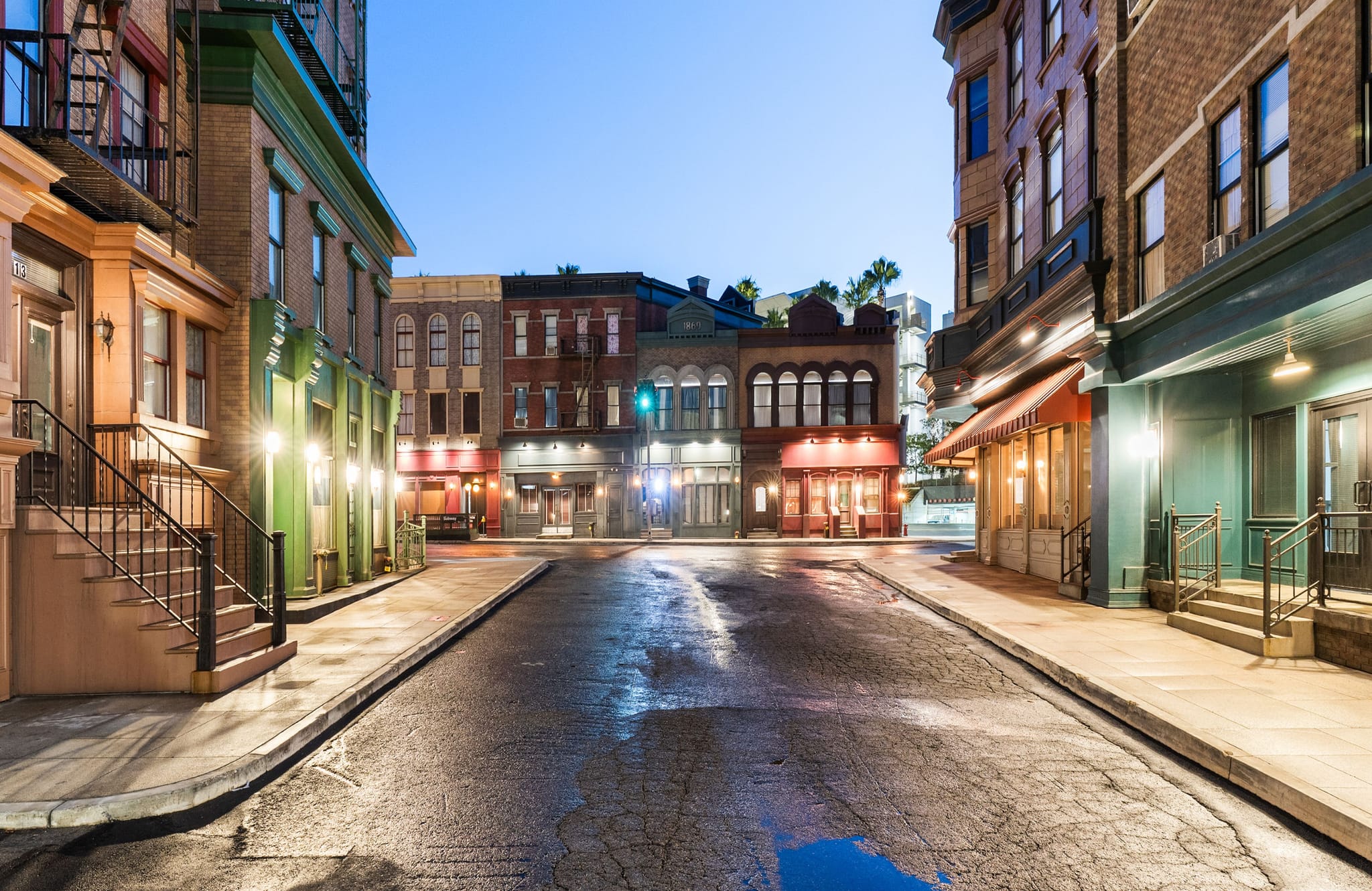 An empty city street at dusk, lined with classic buildings with illuminated windows, reflecting in the wet pavement.