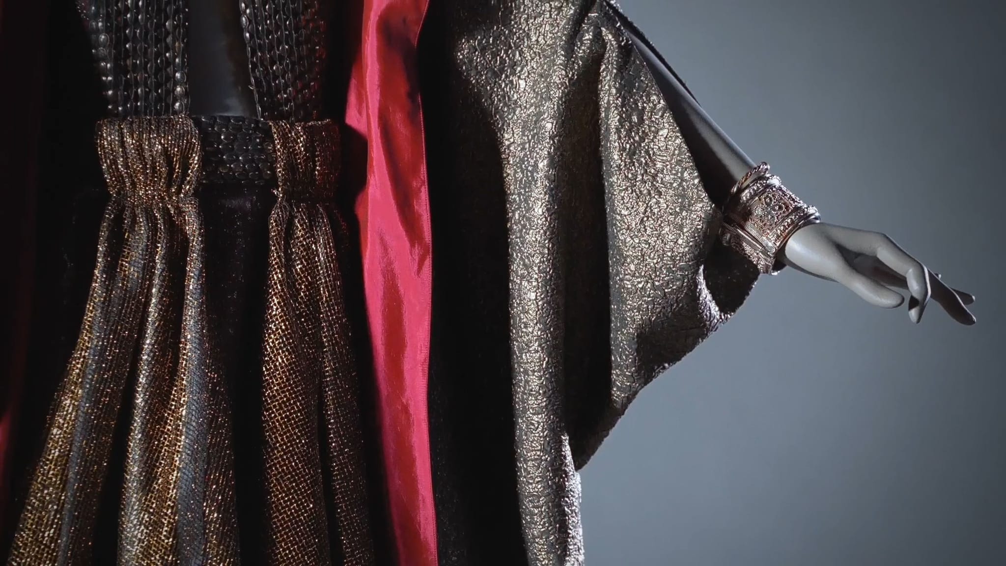 Mannequin wearing a shimmering gold and black gown with a cinched waist, wide sleeves, a red panel, and a cuff bracelet.