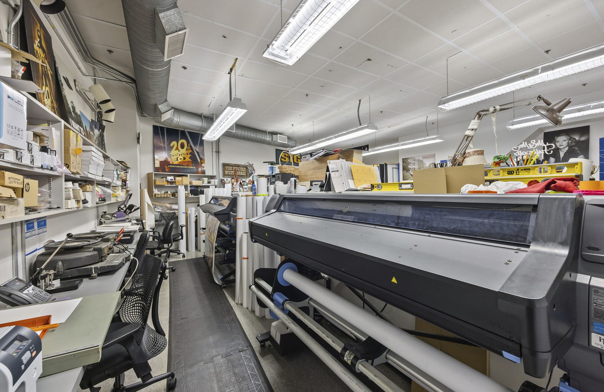 A cluttered print shop with a large format printer, shelves stacked with supplies, and rolls of printing media.