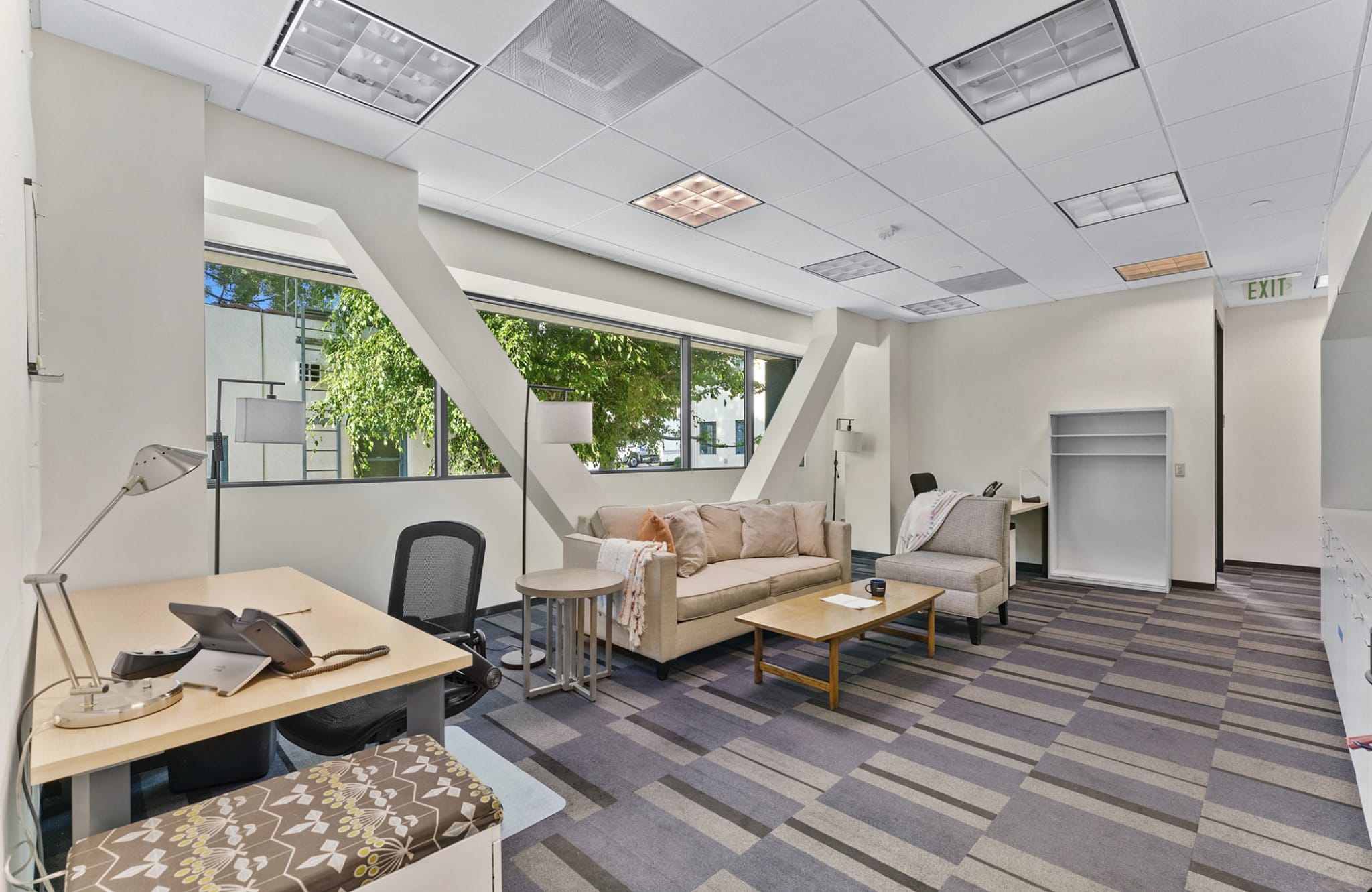 A bright office space with a desk, a lounge area with a beige sofa, large windows, and striped gray carpet.