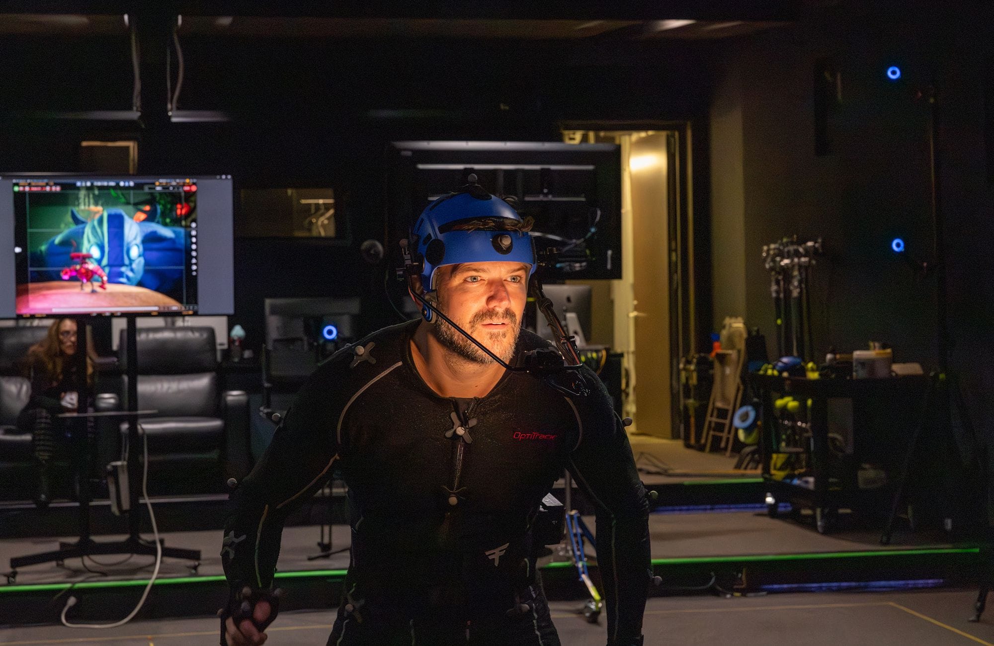 A man in a motion capture suit and helmet performs in a studio, with a monitor displaying a 3D animated character.
