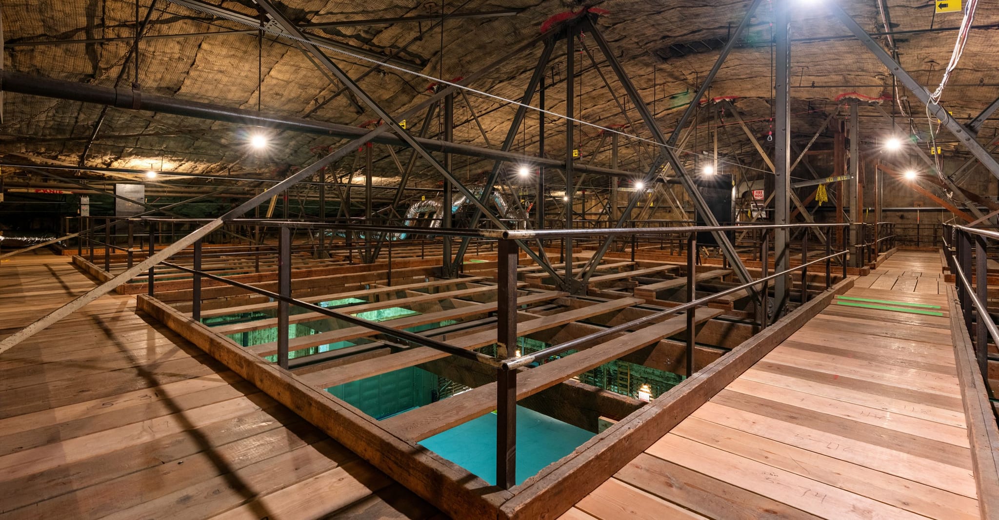 Wooden walkways and metal railings extend over turquoise water in a vast, industrial-style interior with exposed trusses.