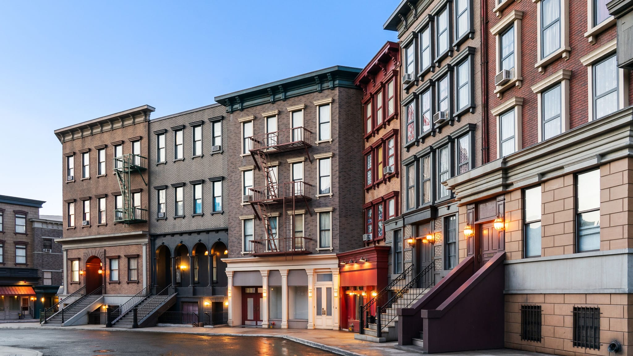 A row of multi-story city buildings with fire escapes and streetlights reflecting on wet pavement at dusk.