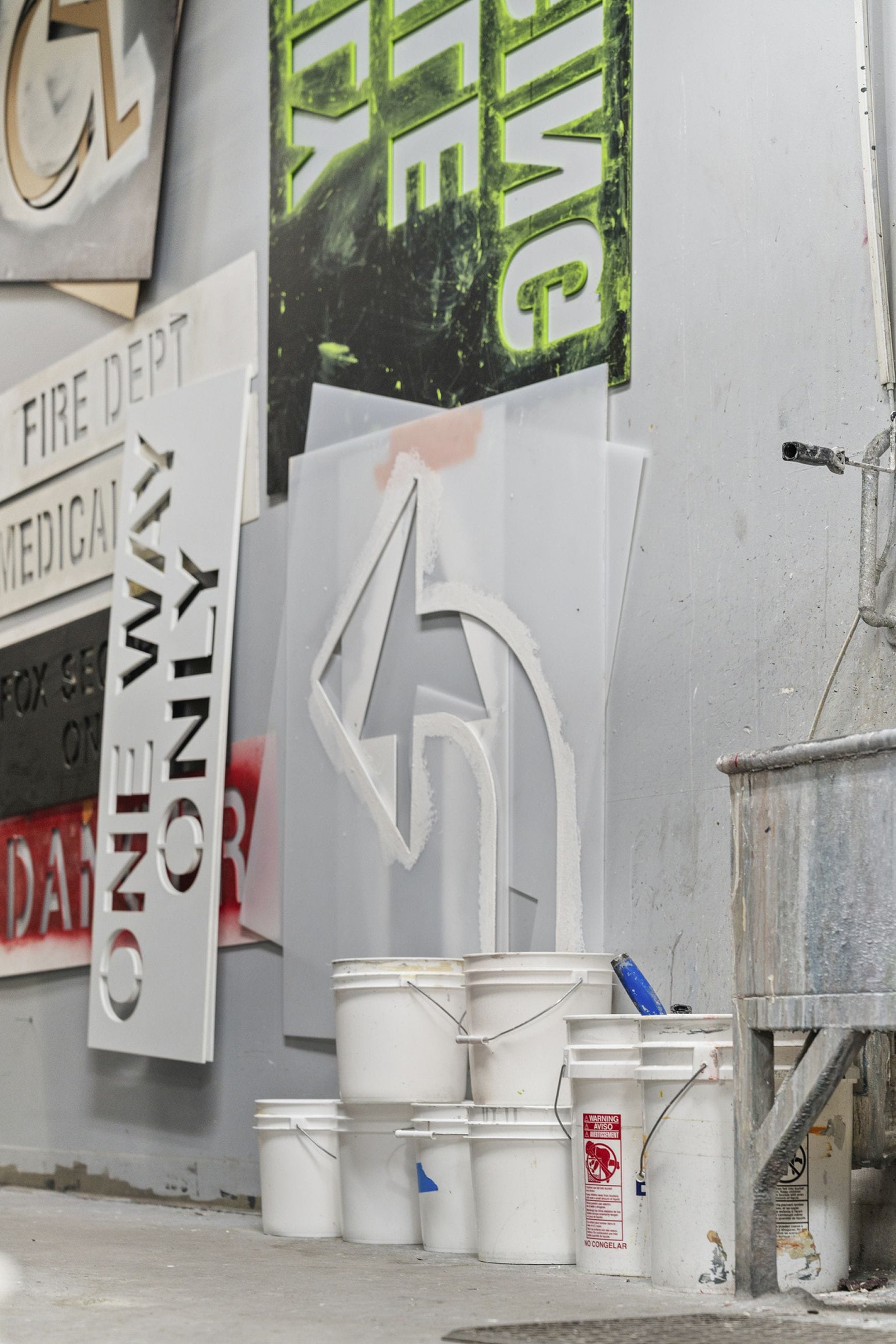 Sign stencils, including a large turn arrow and 'ONE WAY ONLY' text, a green 'SAFETY' sign, and white paint buckets in a workshop.