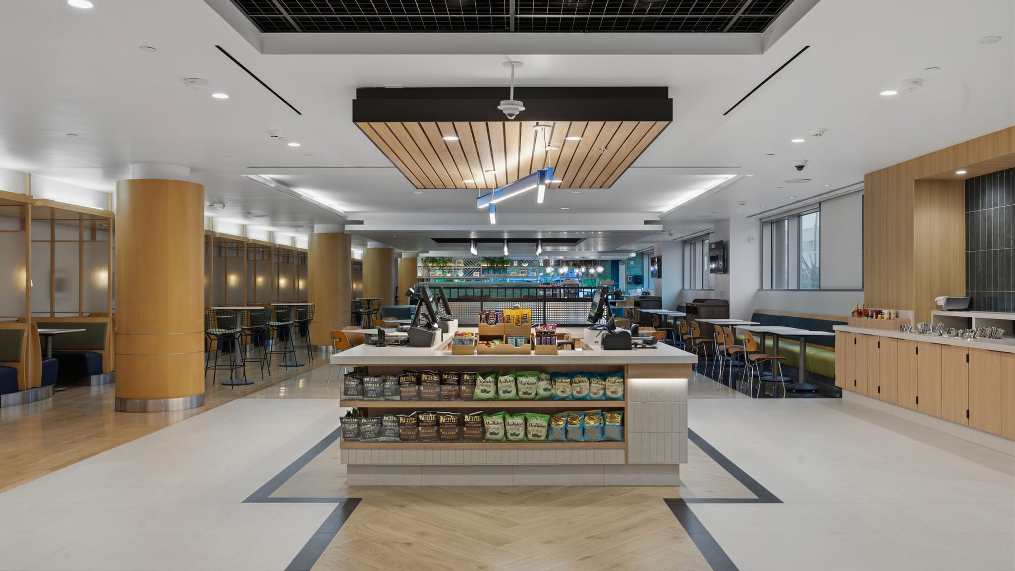 A clean, modern cafeteria or market with a central snack display, various seating areas, and a self-serve station.