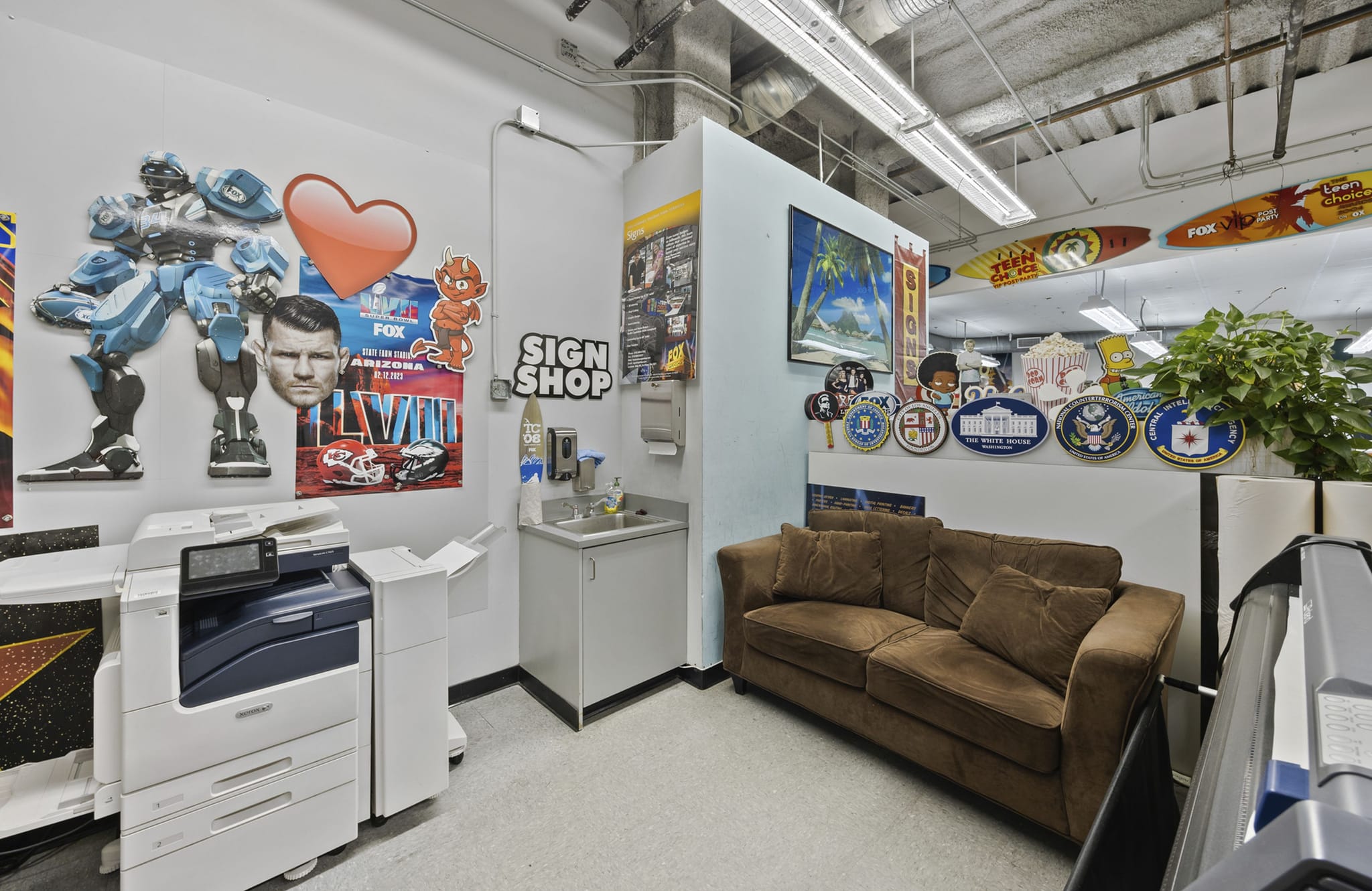 An interior view of a sign shop featuring a large printer, a sink, a brown sofa, and walls decorated with diverse sample signs like a blue robot and Super Bowl poster. Surfboard signs hang from the ceiling.