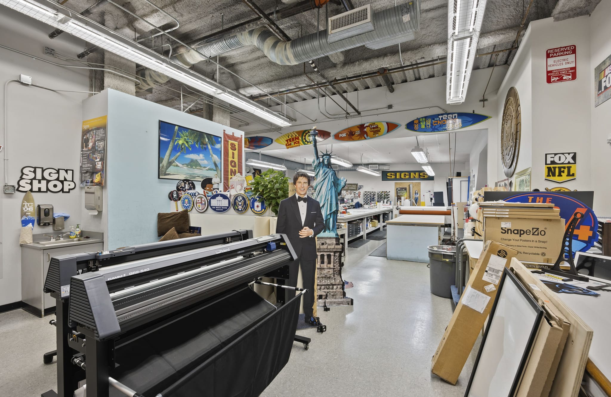 Interior of a lively sign shop, featuring large format printers, numerous sample signs, a tuxedo cutout, and surfboards hanging overhead.
