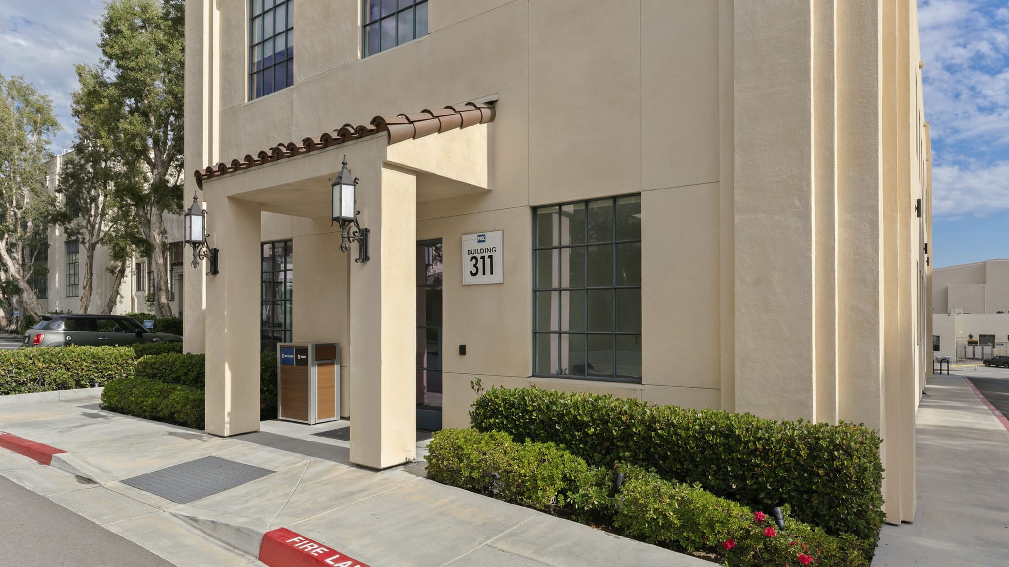 A beige building entrance with a tiled overhang, "BUILDING 311" sign, and green bushes along a sidewalk.