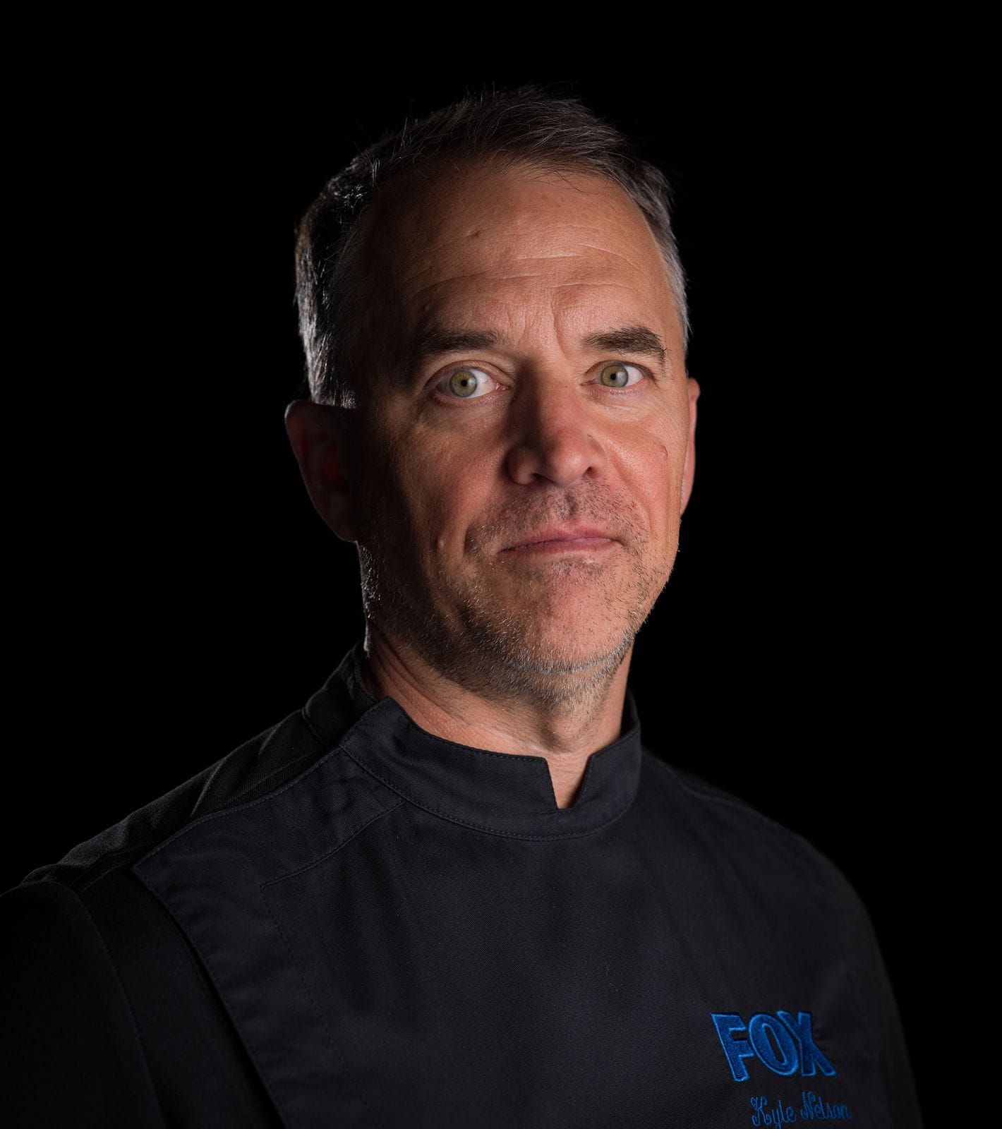 Man with gray hair and light eyes in a black uniform with a blue FOX logo and 'Kyle Nielson' against a black background.