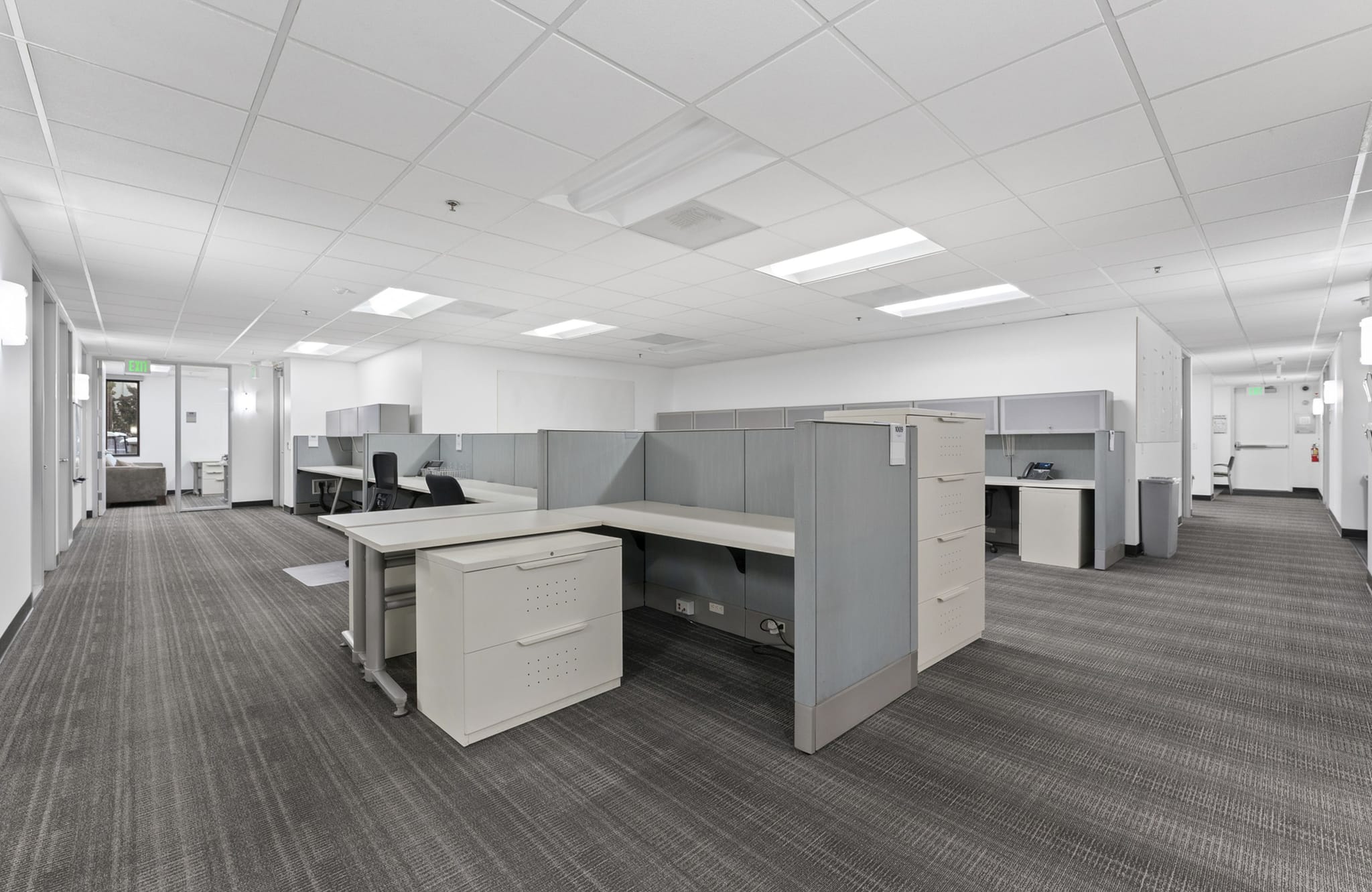 An empty office with gray cubicles, gray patterned carpet, and bright overhead lighting.