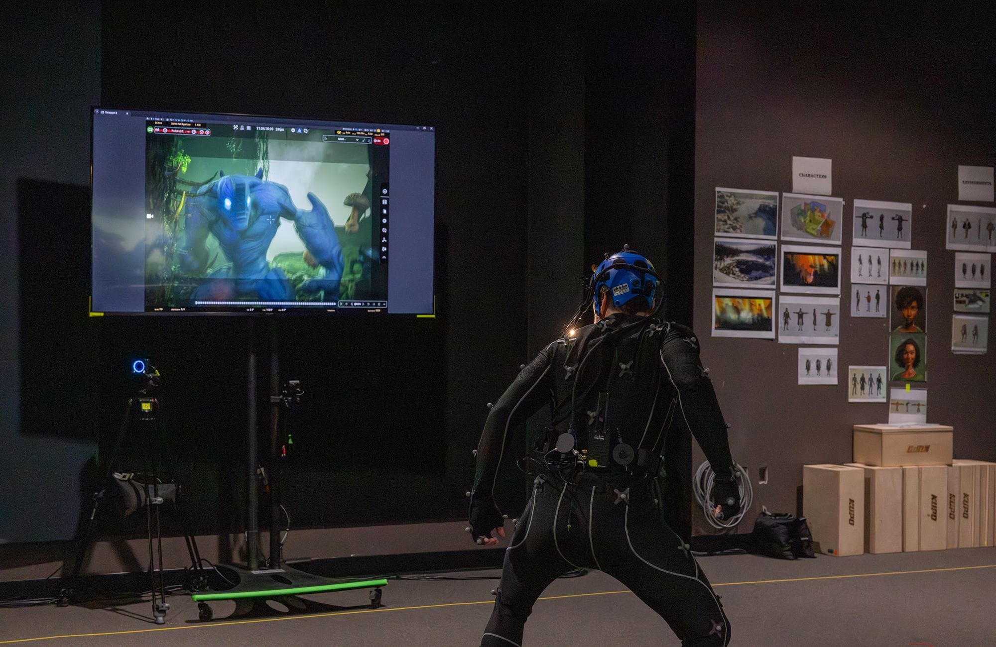 A person in a motion capture suit performs in a studio, facing a large screen displaying a blue animated character, with concept art on the wall.