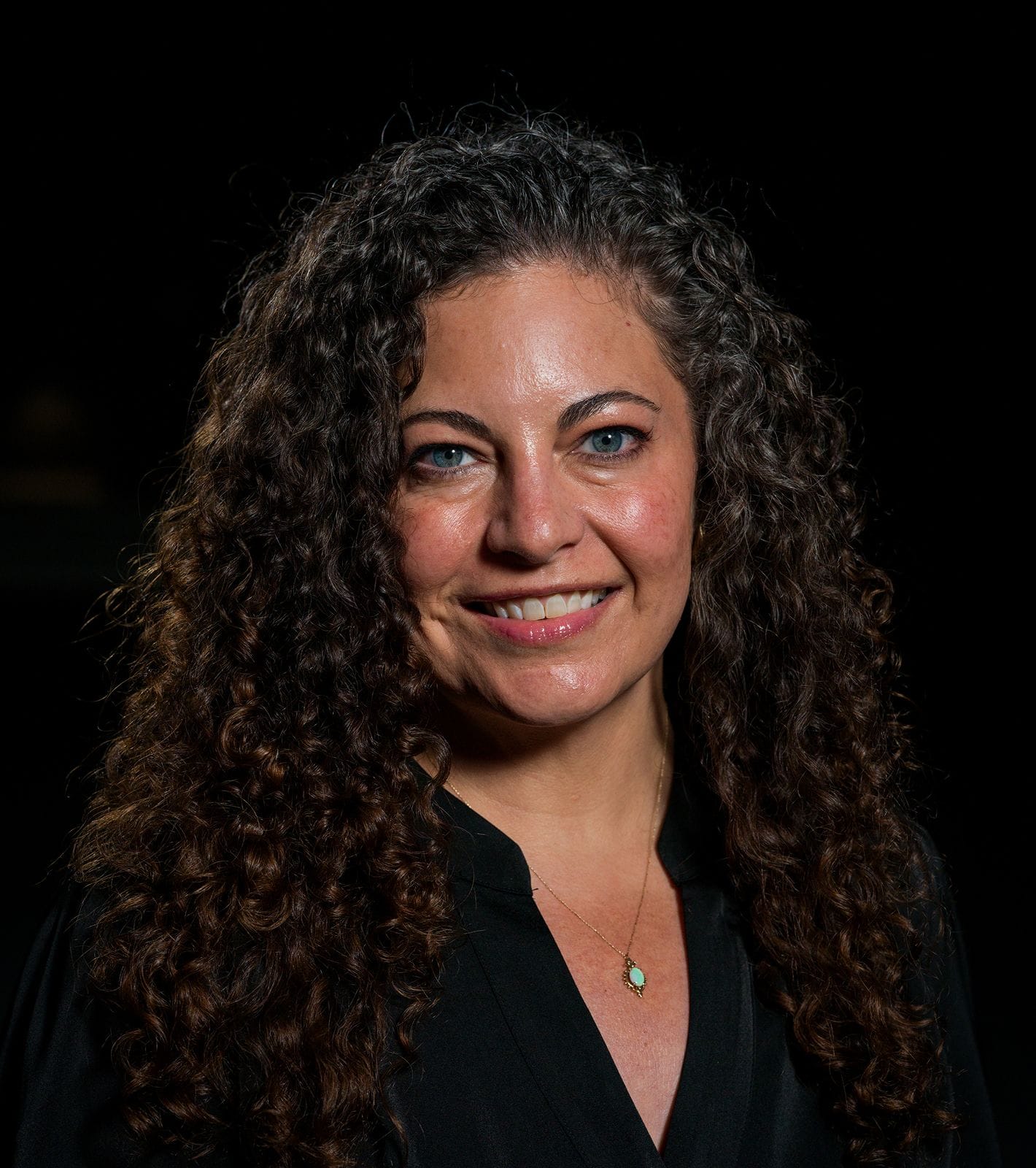 A smiling woman with long curly brown hair and blue eyes wears a black shirt against a dark background.