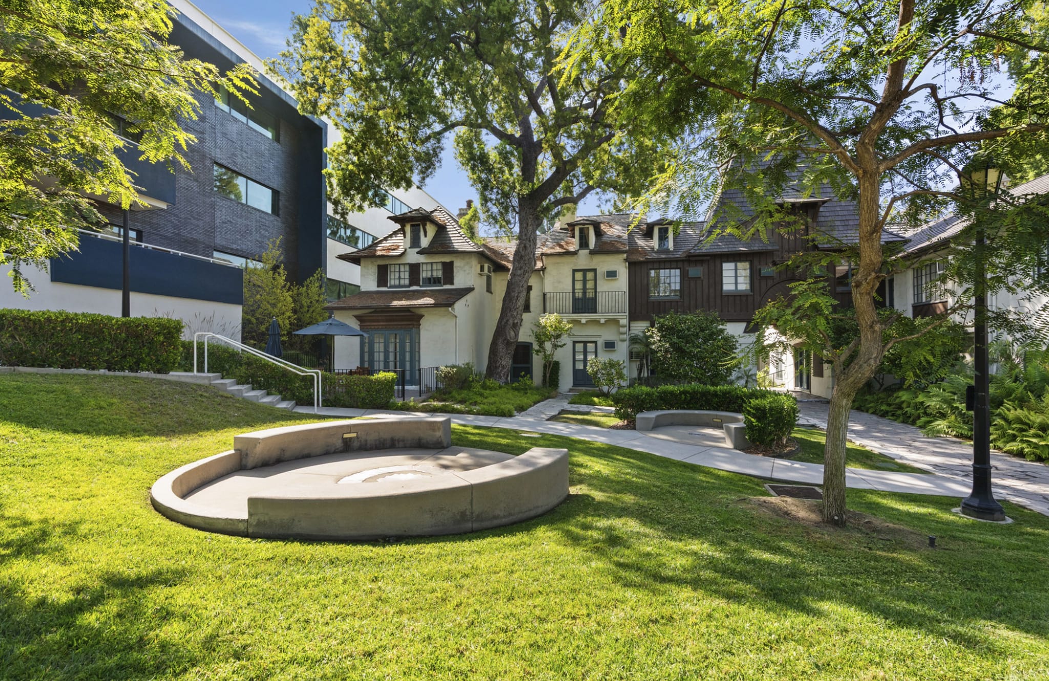 A landscaped outdoor area featuring a modern dark building on the left and several traditional, multi-story buildings on the right, separated by a green lawn with a curved concrete seating area and mature trees.
