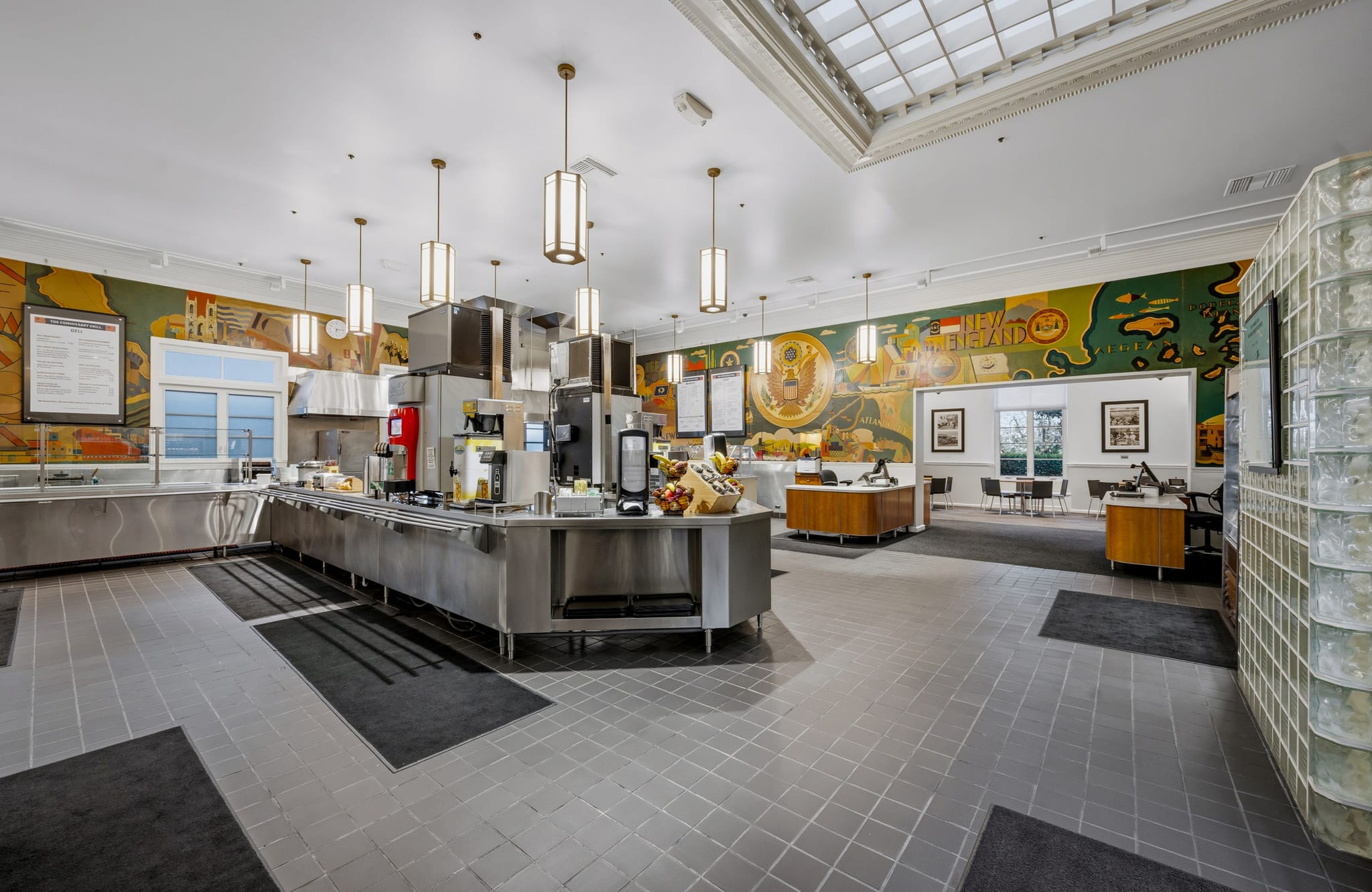 A spacious cafeteria with stainless steel serving counters, colorful murals, a skylight, and a glass block wall.