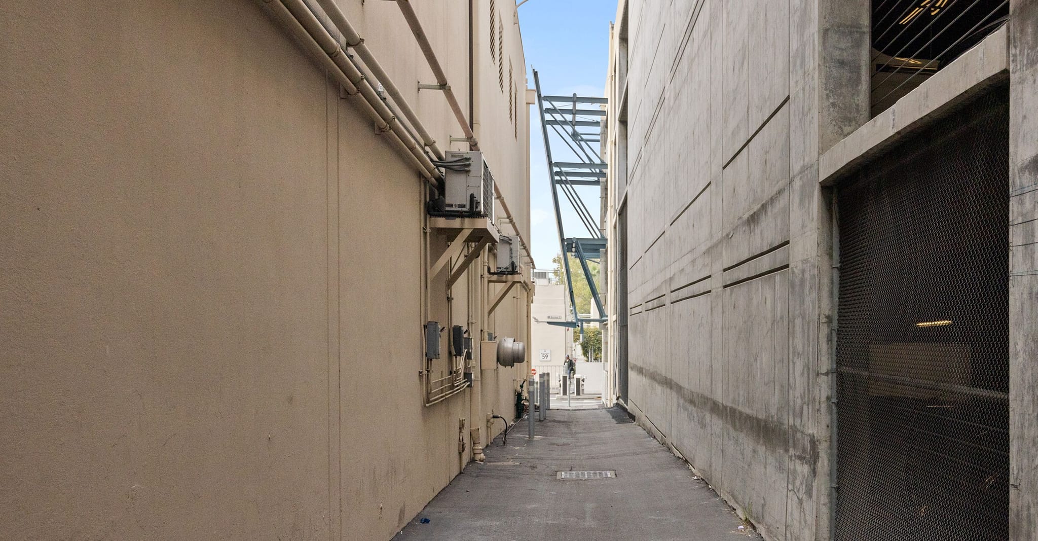 A narrow alleyway between a tan building and a concrete building, with structural elements visible in the distance.