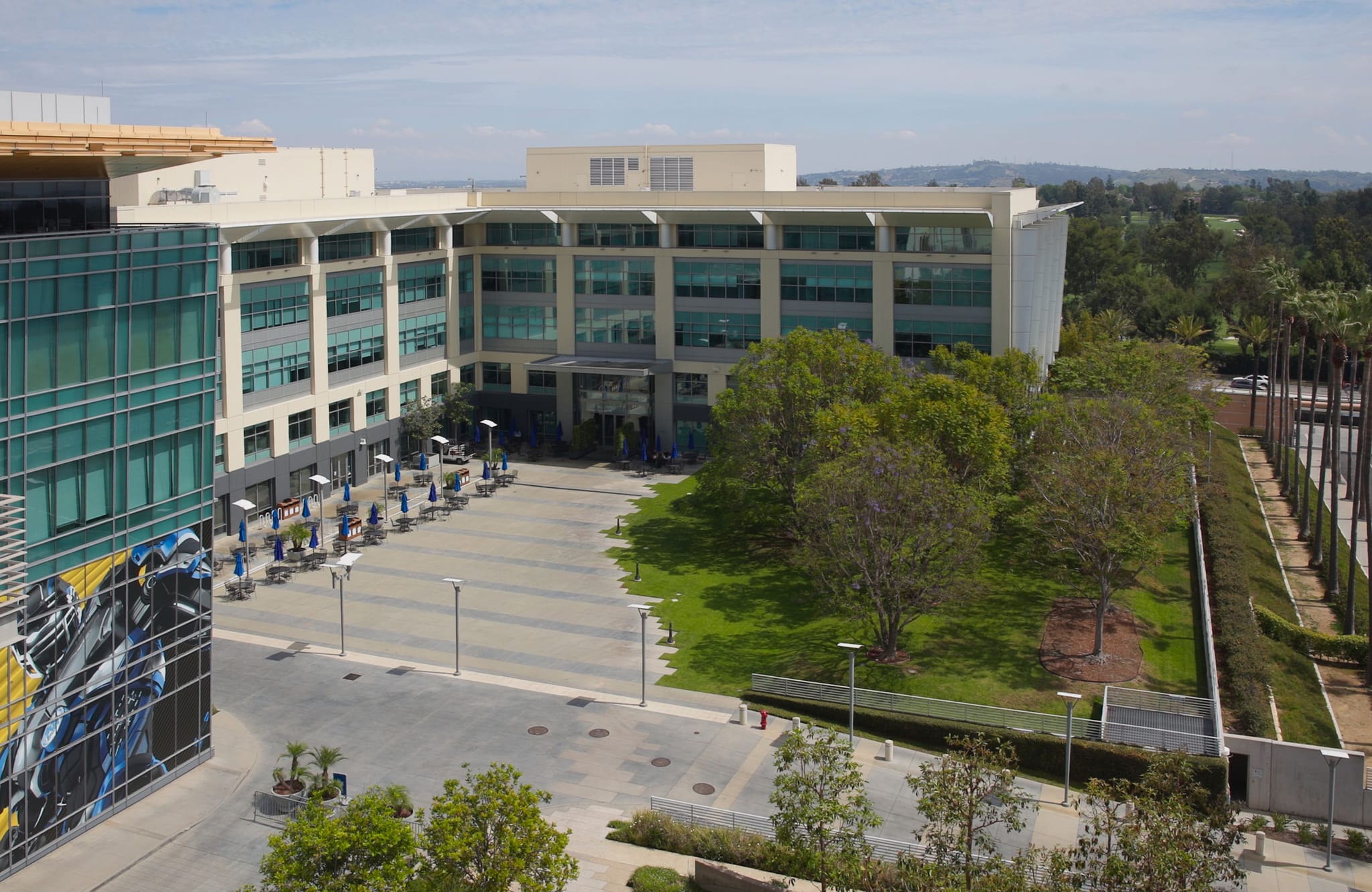 A modern office campus with large buildings, an outdoor plaza with many tables and blue umbrellas, green lawns, and distant hills.