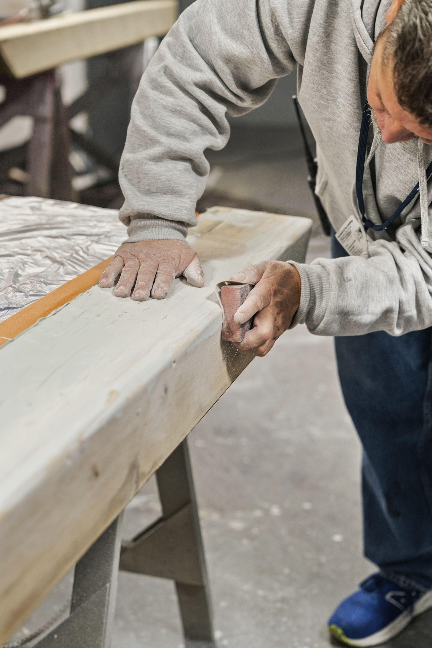 A person in a grey hoodie sands a wooden beam with a sanding block.