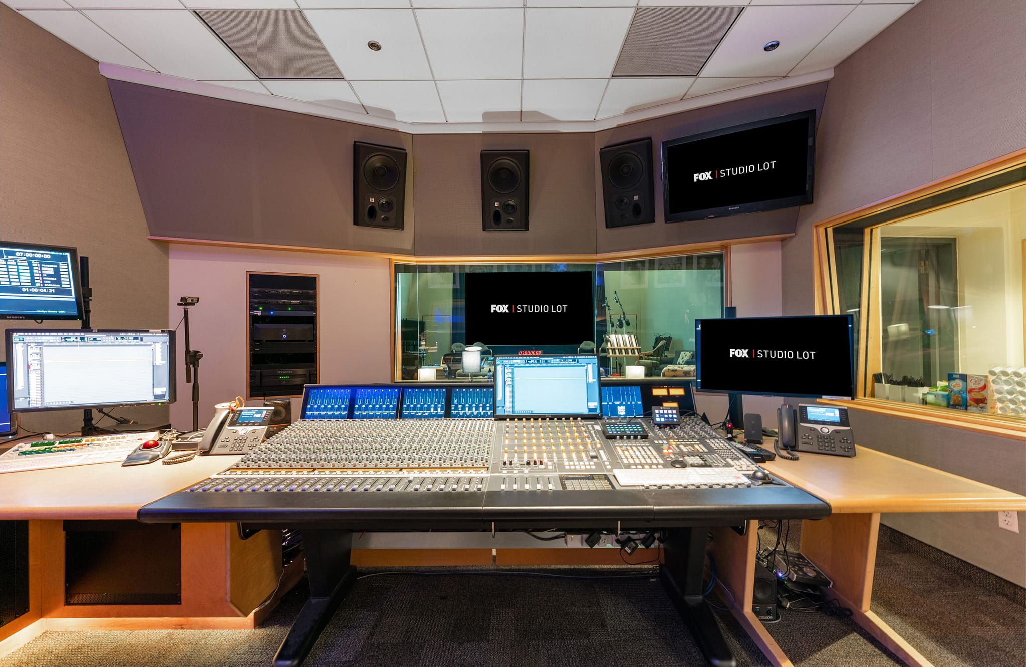 A professional audio-video control room with a large mixing console, multiple monitors, and screens displaying 'FOX STUDIO LOT'.