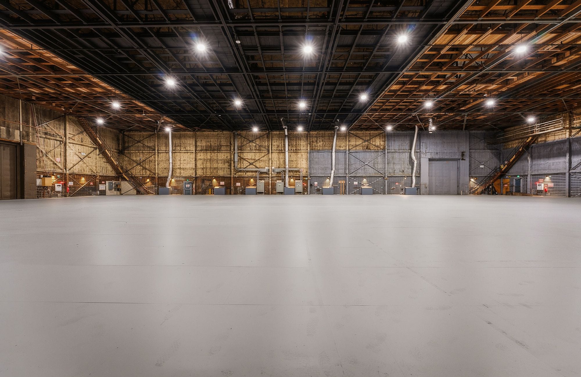 A large, empty studio space with a light gray floor, exposed beam ceiling, and bright overhead lights.